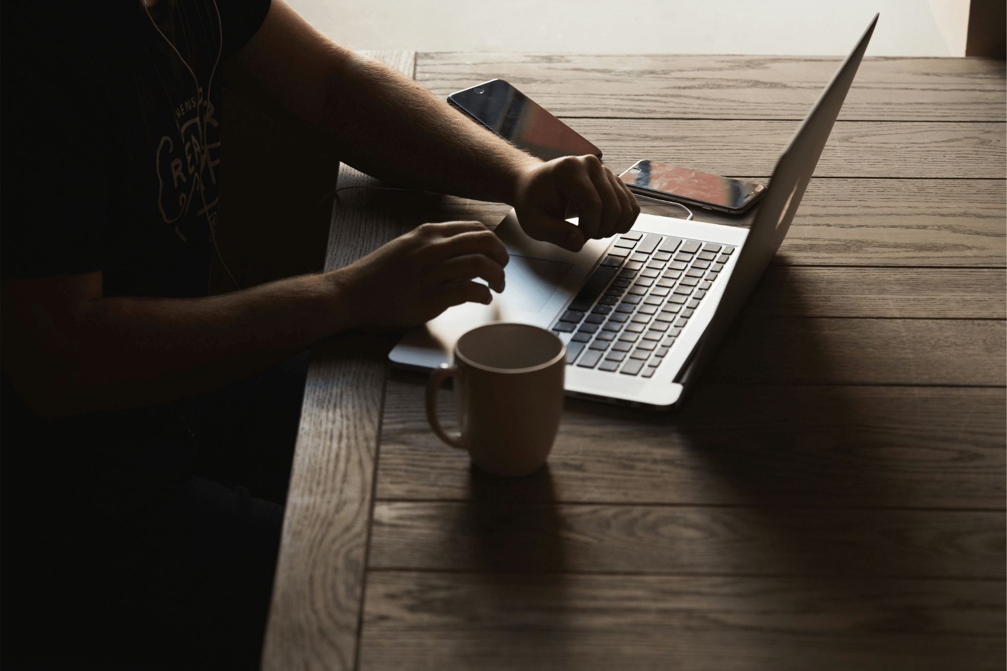 man at table typing on a laptop with a coffee mug and mobile devices nearby