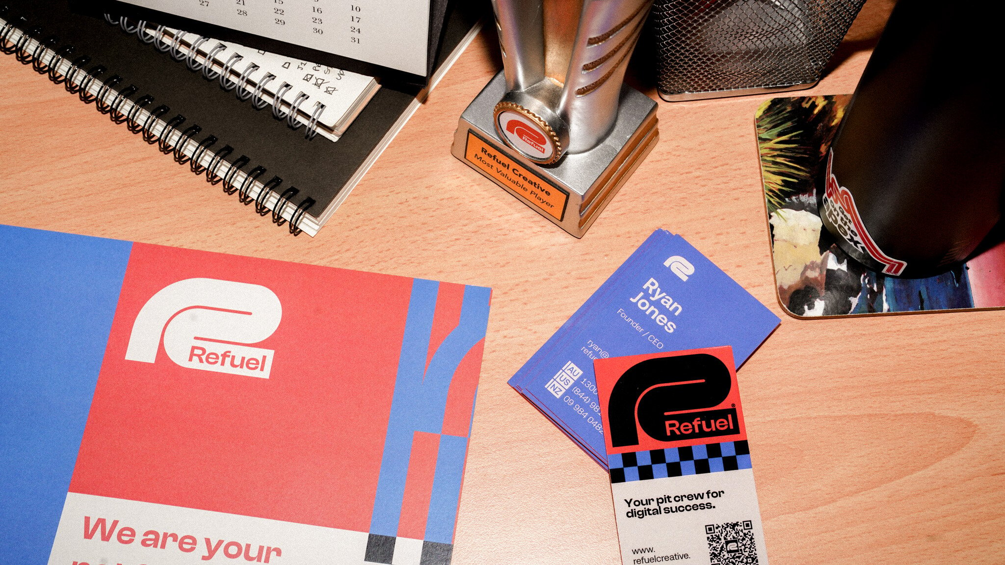 Refuel Creative branded marketing materials spread on a wooden desk, including business cards, a notebook, and what appears to be a trophy or award. The Refuel logo is prominently displayed in red and blue colours.