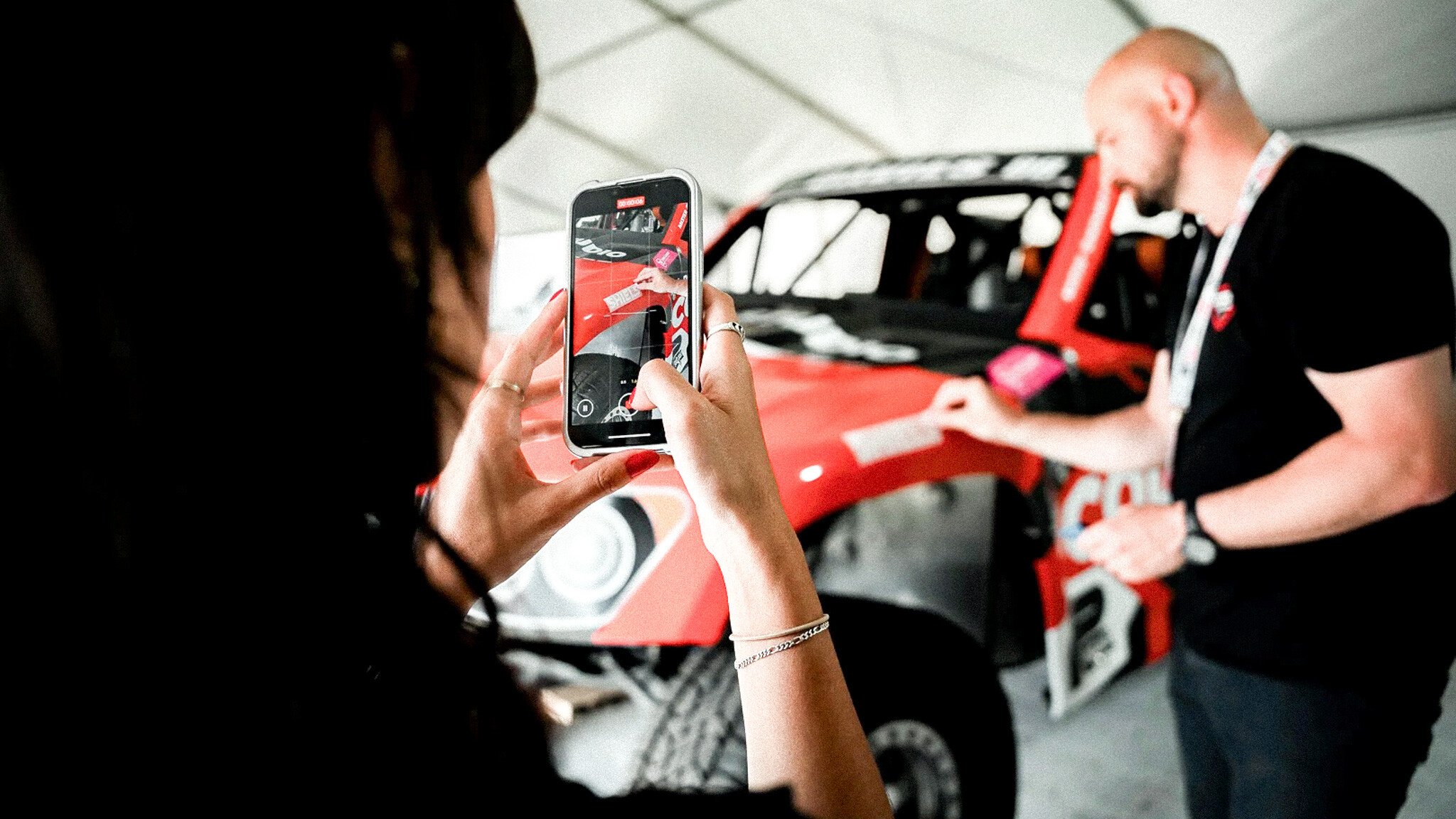 Over-the-shoulder view of someone filming a video with their phone of a red super truck and a person in a garage.