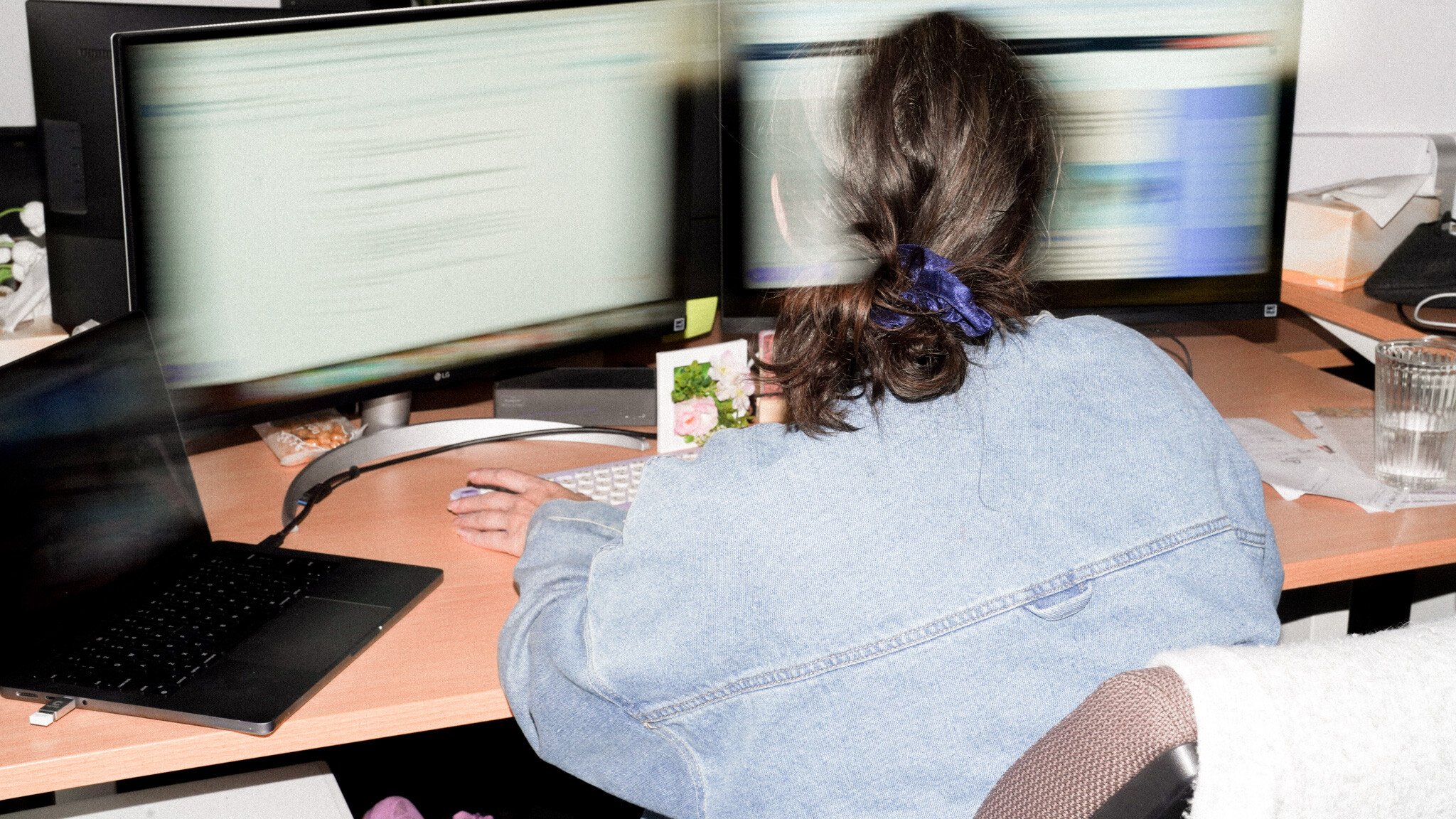 Rear view of someone with a blue hair tie and denim jacket working at a desk with multiple monitors, showing motion-blurred screens.