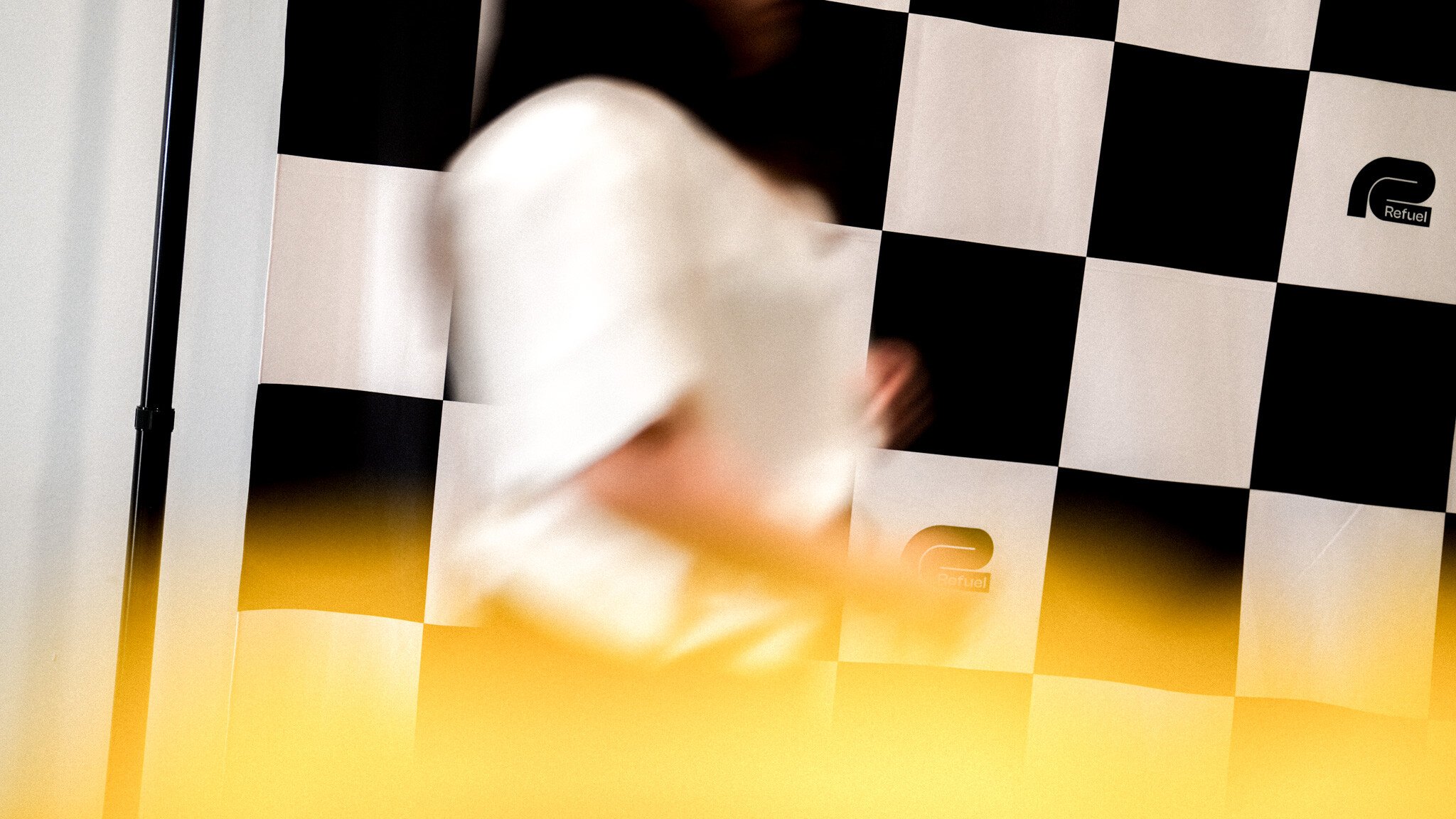 Abstract blurred motion image featuring a black and white checkered pattern background with a yellow gradient at the bottom. A white blurred shape is visible in the center.