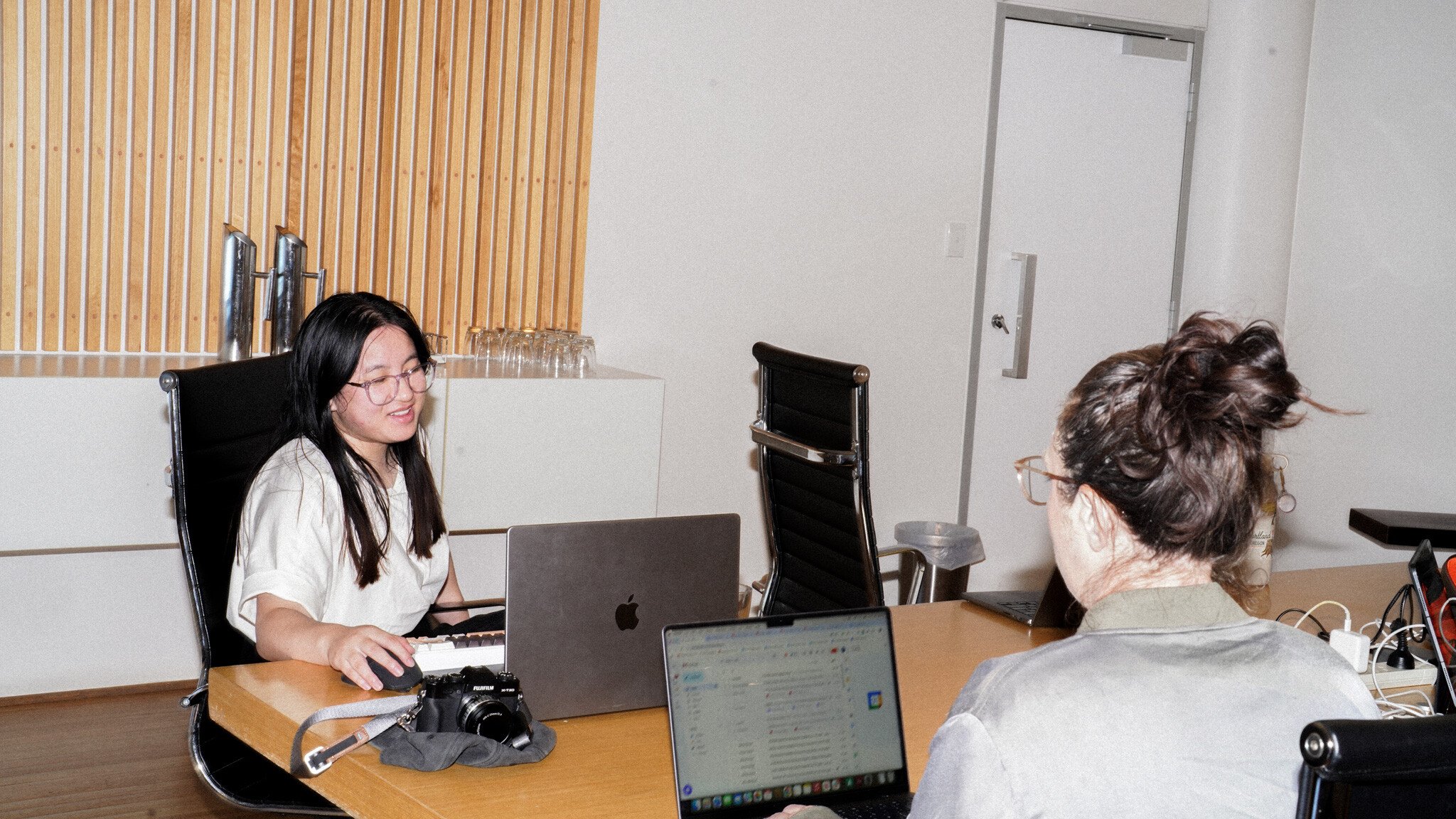 Two people at a wooden table having a meeting, both working on MacBooks. They're sitting across from each other in black office chairs, with wooden slat wall decor visible in the background.