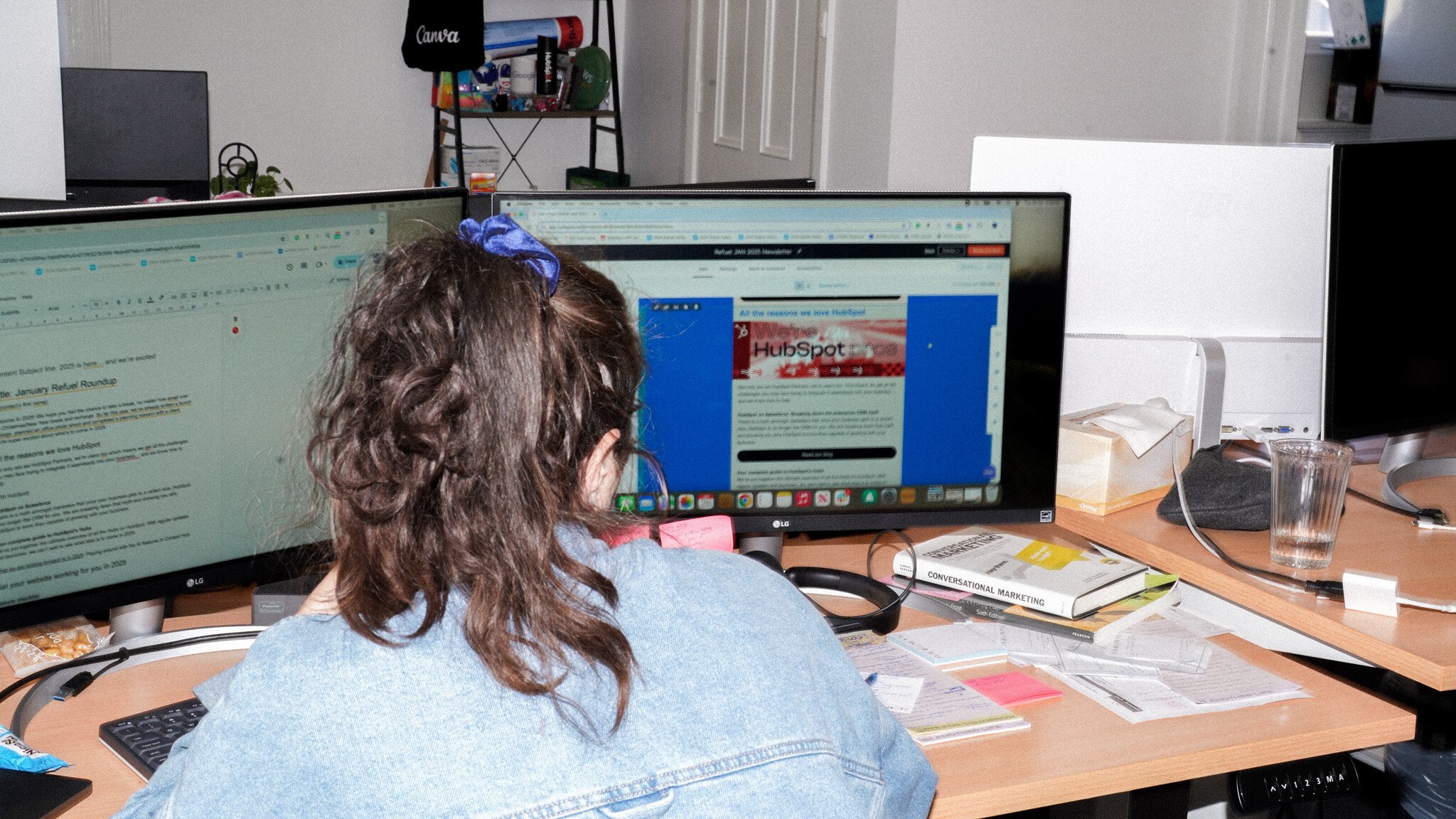 Rear view of someone with a blue hair tie wearing a denim shirt, working at a desk with dual monitors displaying documents and web content. Office supplies and papers are scattered on the desk.