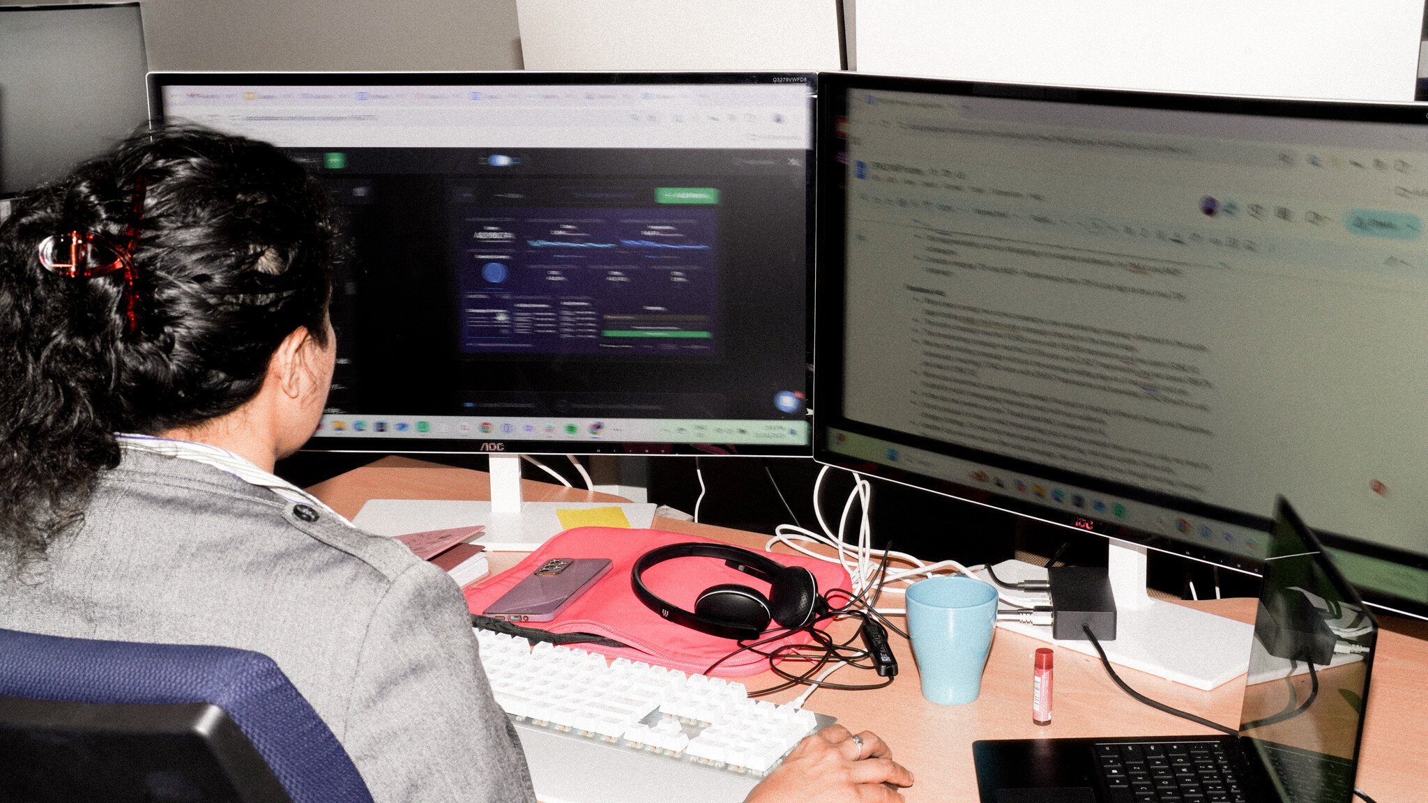 Over-the-shoulder view of someone working at a dual-monitor setup, with one screen showing a dark-themed dashboard with data visualisations and the other displaying text content. A headset, blue mug, and other desk accessories are visible.