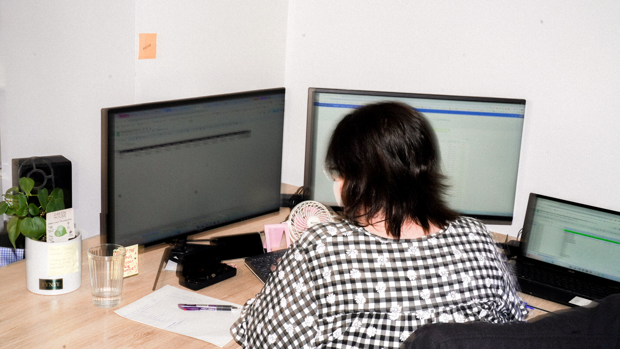 Rear view of someone in a checkered shirt working at a desk with multiple monitors and a laptop, all displaying spreadsheets or data. A plant and glass are visible on the desk's corner.