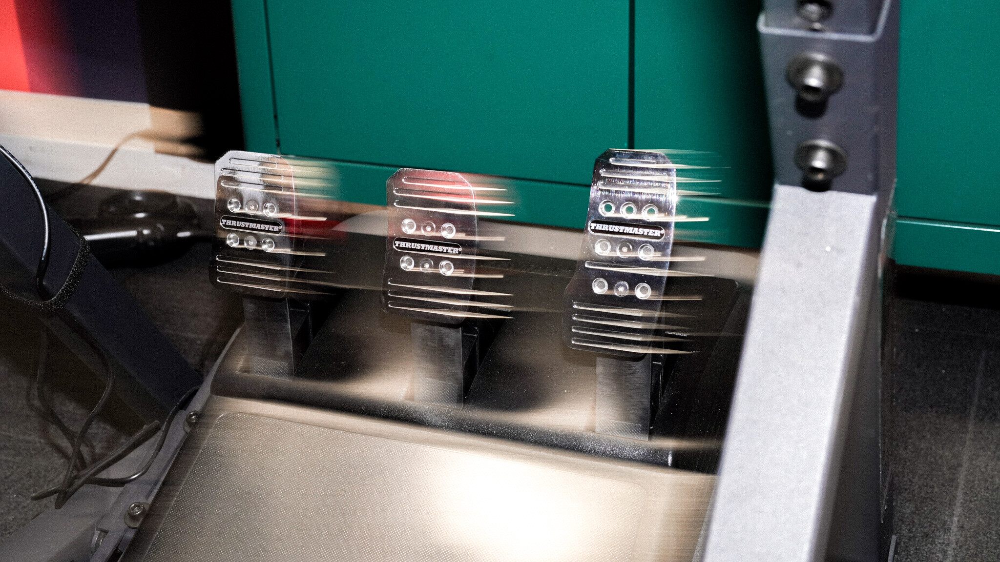 Motion-blurred shot of three racing pedals (likely gas, brake, and clutch) mounted on a metallic platform against a green background.