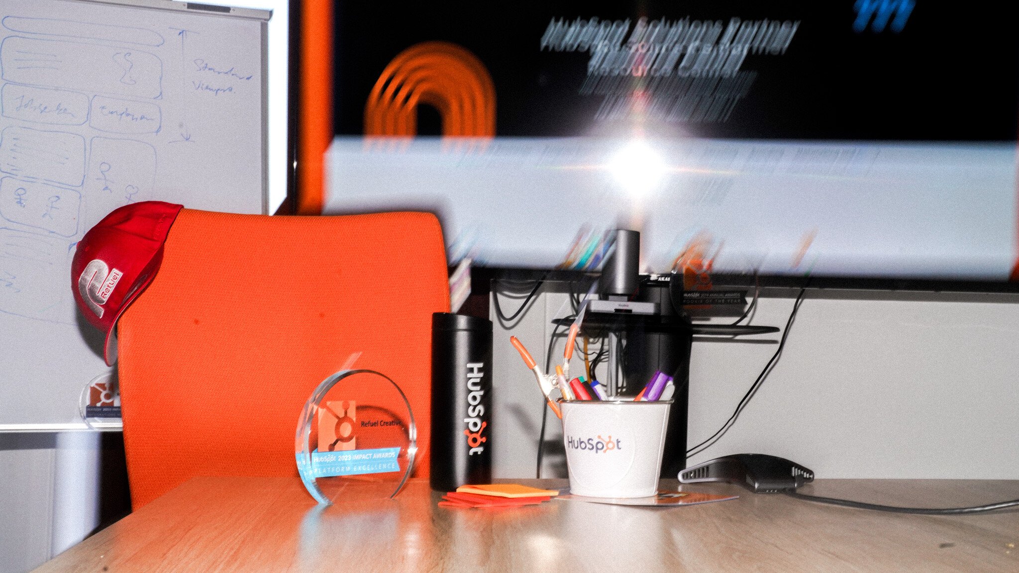 A desk with Refuel Creative and HubSpot branded items including an award trophy, cap, water bottle, and pen holder. A monitor displaying a presentation is visible in the background.