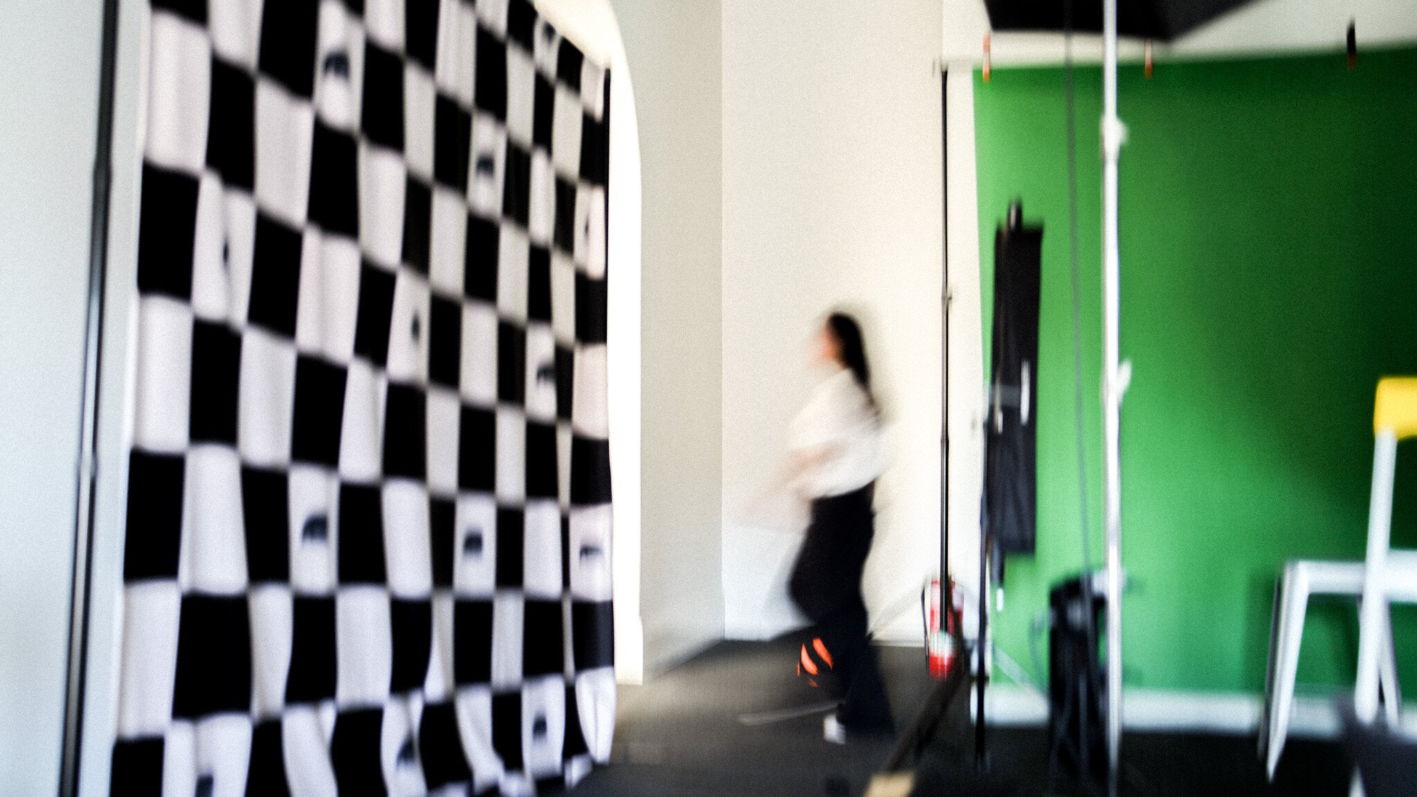 Motion-blurred shot of a studio setup with a black and white checkered backdrop on the left, a blurred figure in white and black moving past, and a green screen visible in the background.