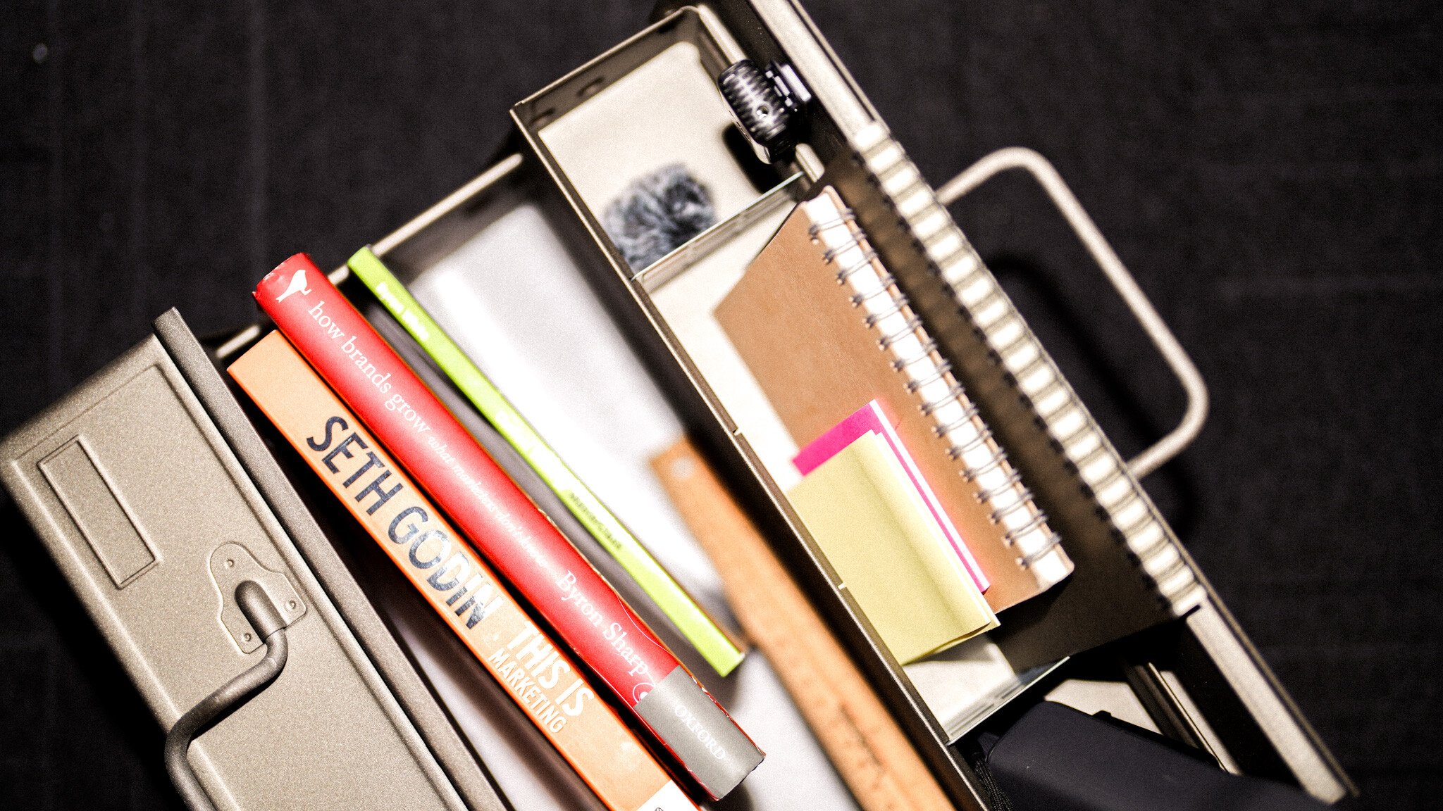 Angled view of business books including Seth Godin's work and 'How Brands Grow' alongside a metal organiser containing notebooks and sticky notes.