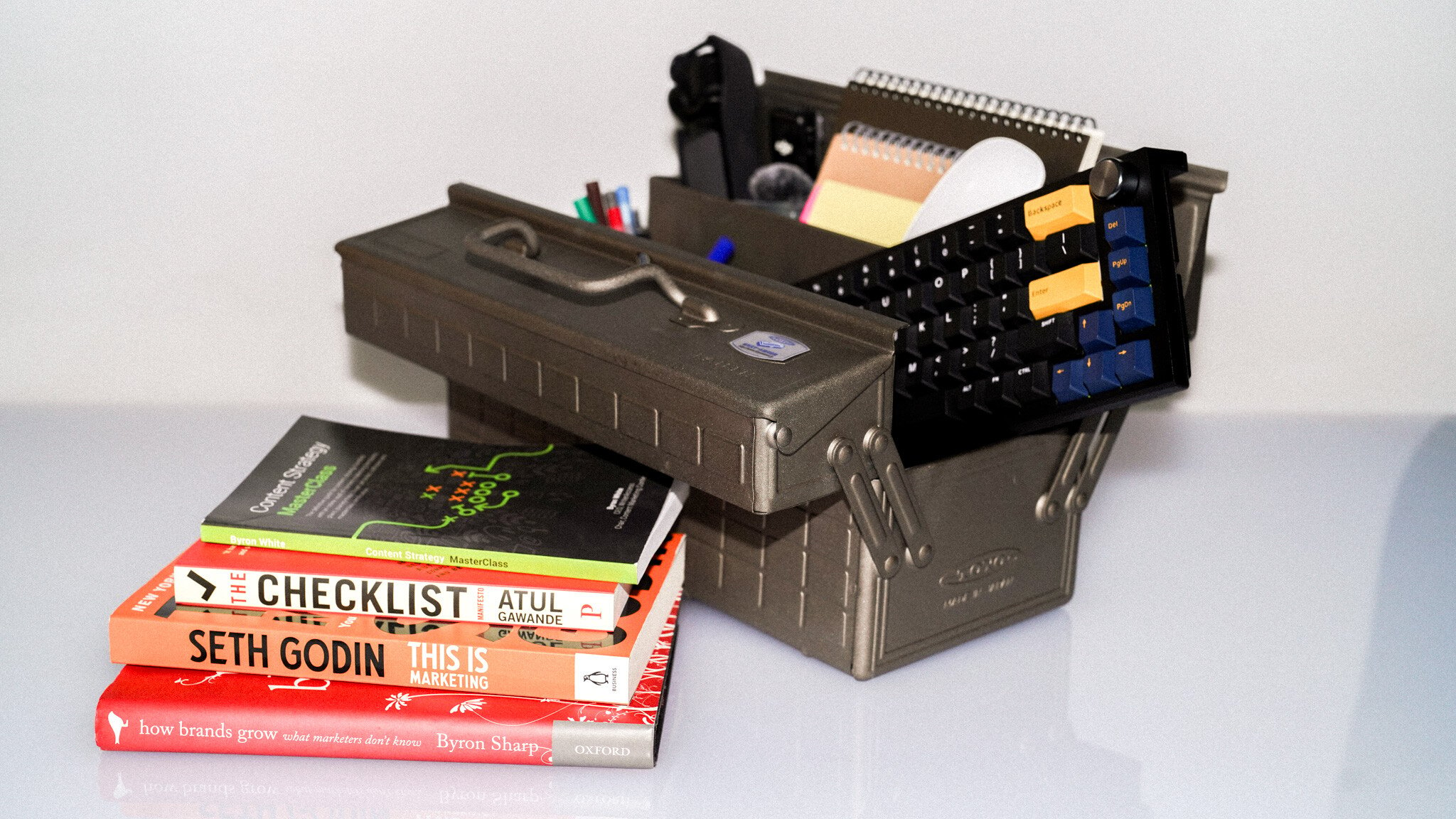 A stack of business books including 'Checklist' by Atul and a Seth Godin book, next to a dark metal organiser containing office supplies and keyboards.