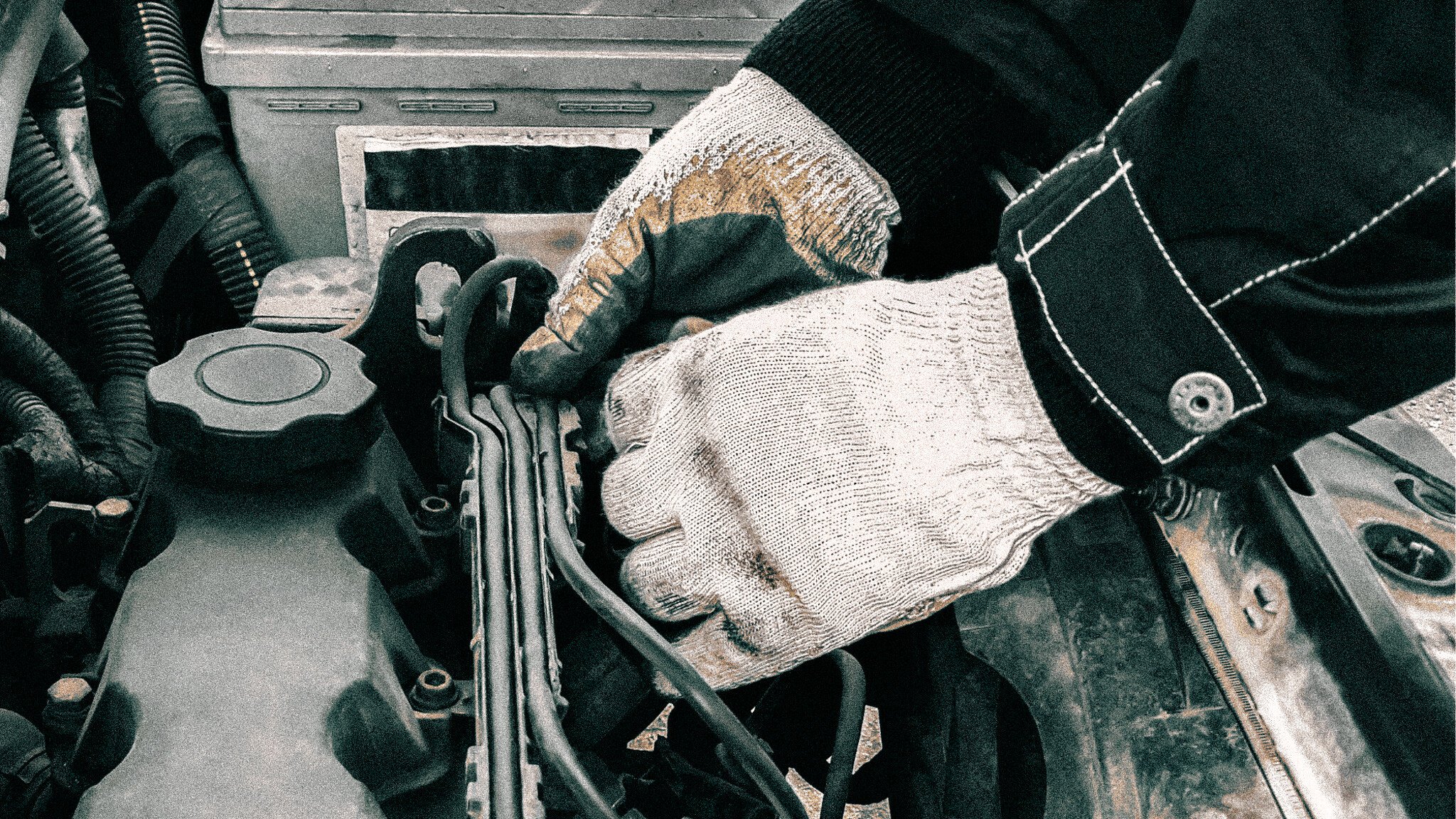 Close-up of hands wearing dirty work gloves reaching into a car engine compartment, with various mechanical components and an oil cap visible.