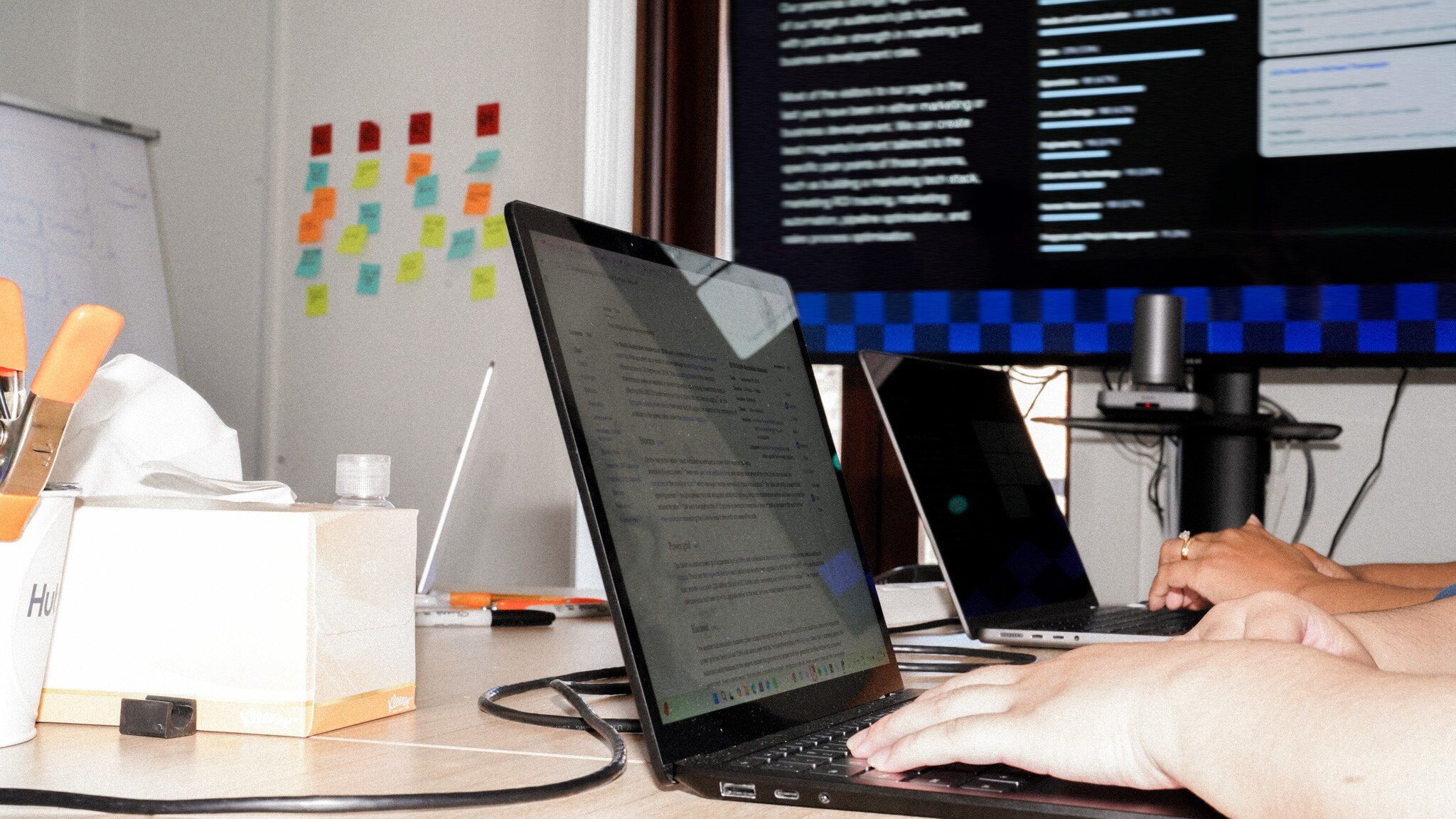 Workspace showing hands typing on a laptop, with a wall of colourful sticky notes in the background and a monitor displaying code with a blue checkered pattern at the top.