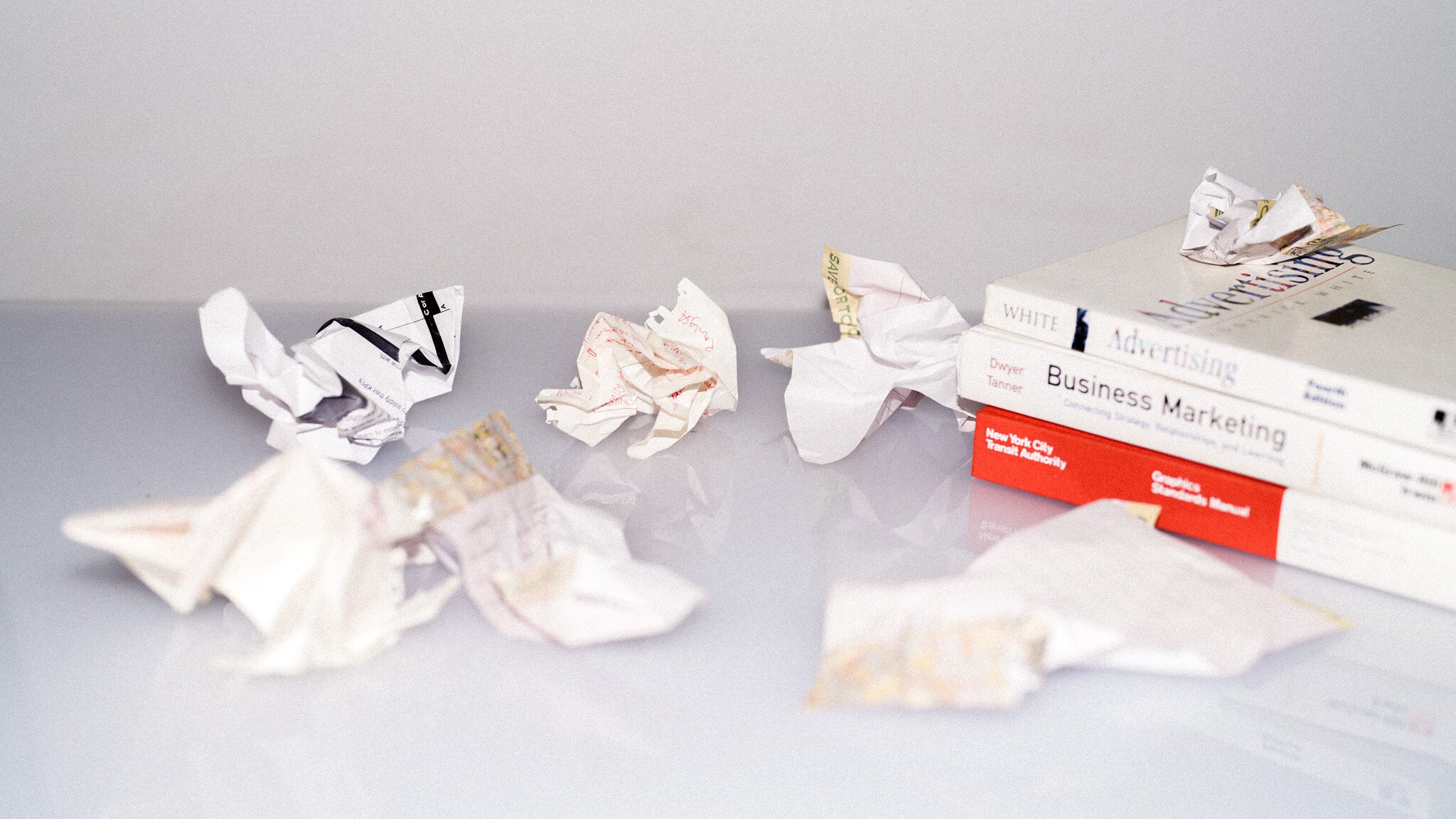 Crumpled paper balls scattered on a white surface next to business marketing textbooks, suggesting frustration or discarded ideas during study or work.