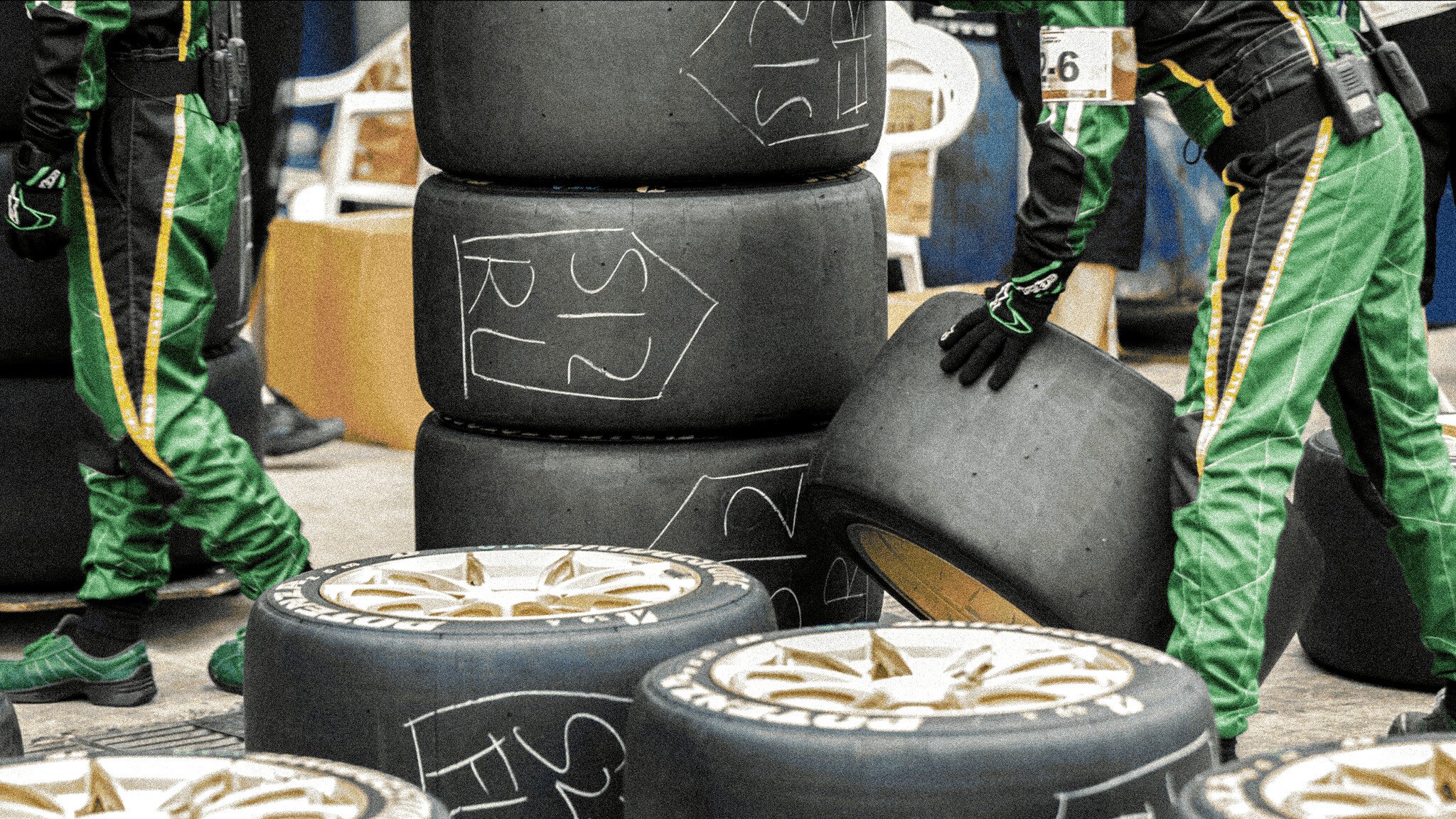 Racing tires stacked on the ground with numbers written in chalk on their sides. Gold-coloured wheel rims are visible, and people wearing green racing suits are standing nearby.