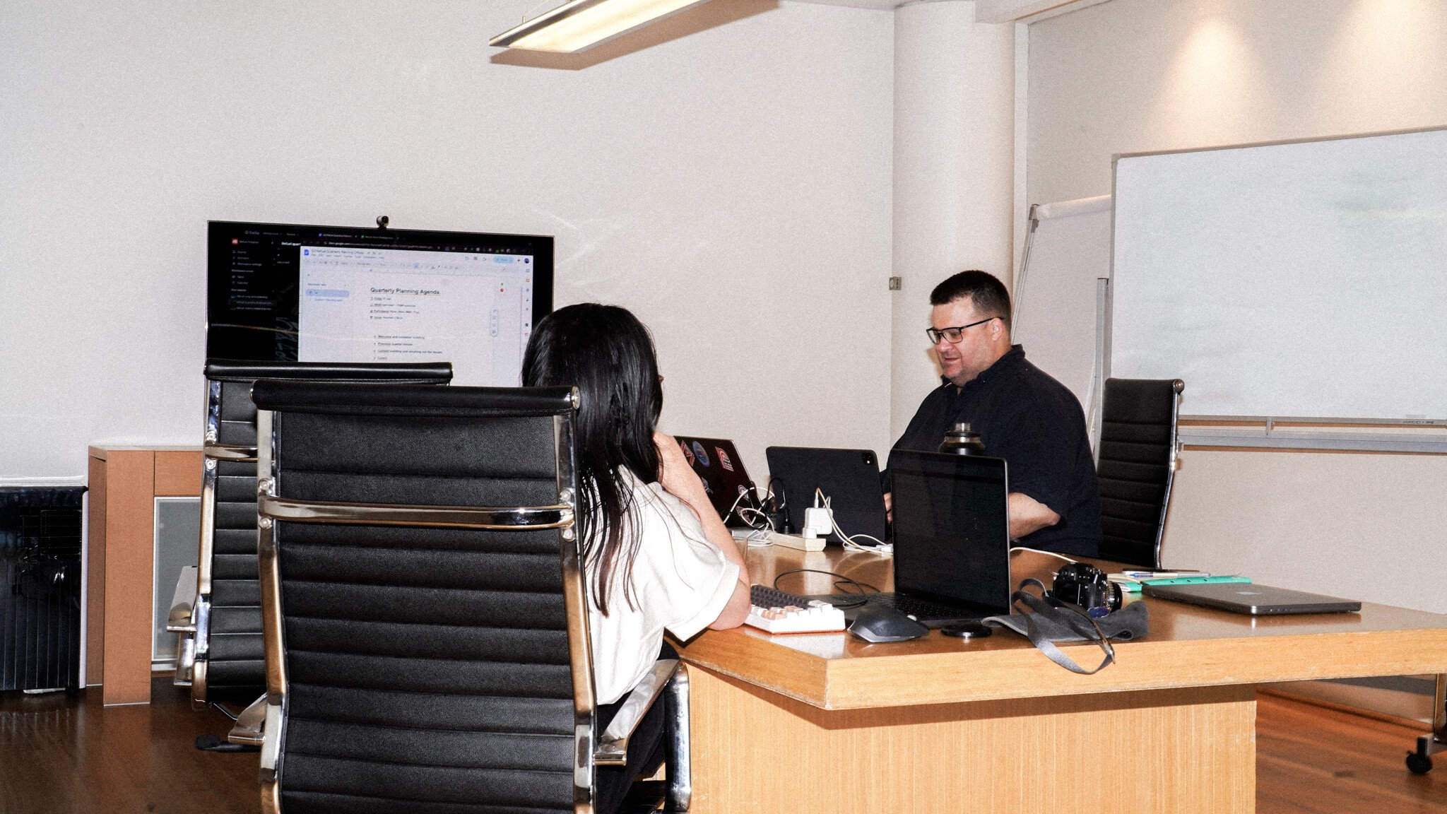 A meeting room showing two people at a wooden desk with laptops and a monitor displaying an agenda. Black office chairs and a whiteboard are visible in the room.