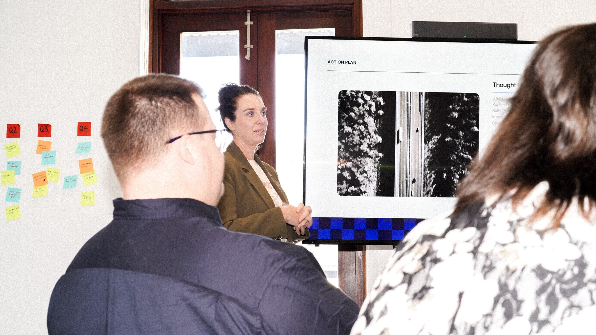 A presentation in progress showing someone in a brown blazer discussing content on a screen displaying 'Action Plan' and images. Colourful sticky notes are visible on the wall and attendees are viewing the presentation.