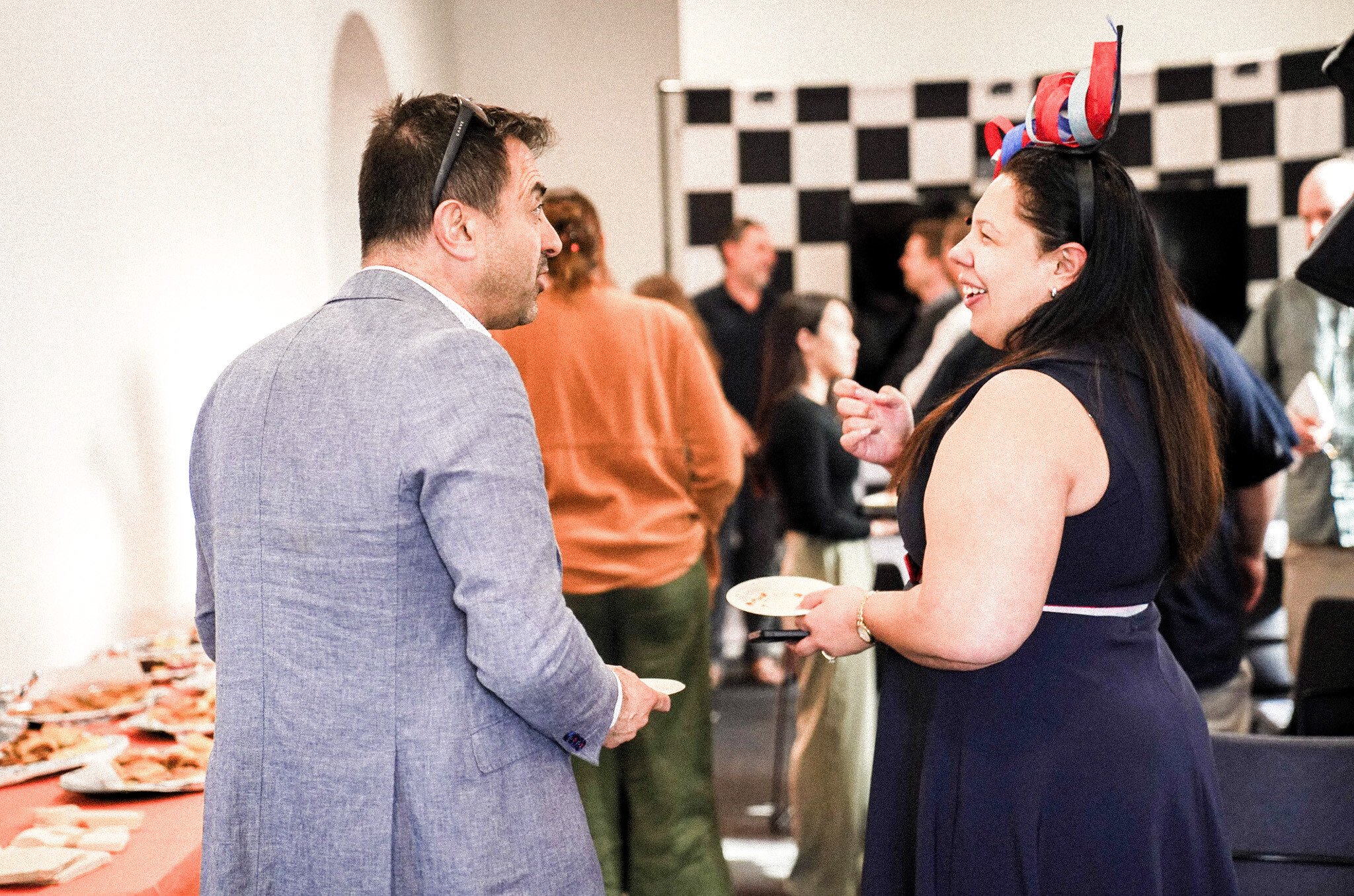 Two people engaged in conversation at a social gathering, one wearing a grey suit jacket and another in a navy dress with a red and blue headpiece. A checkered pattern wall and buffet table are visible in the background.