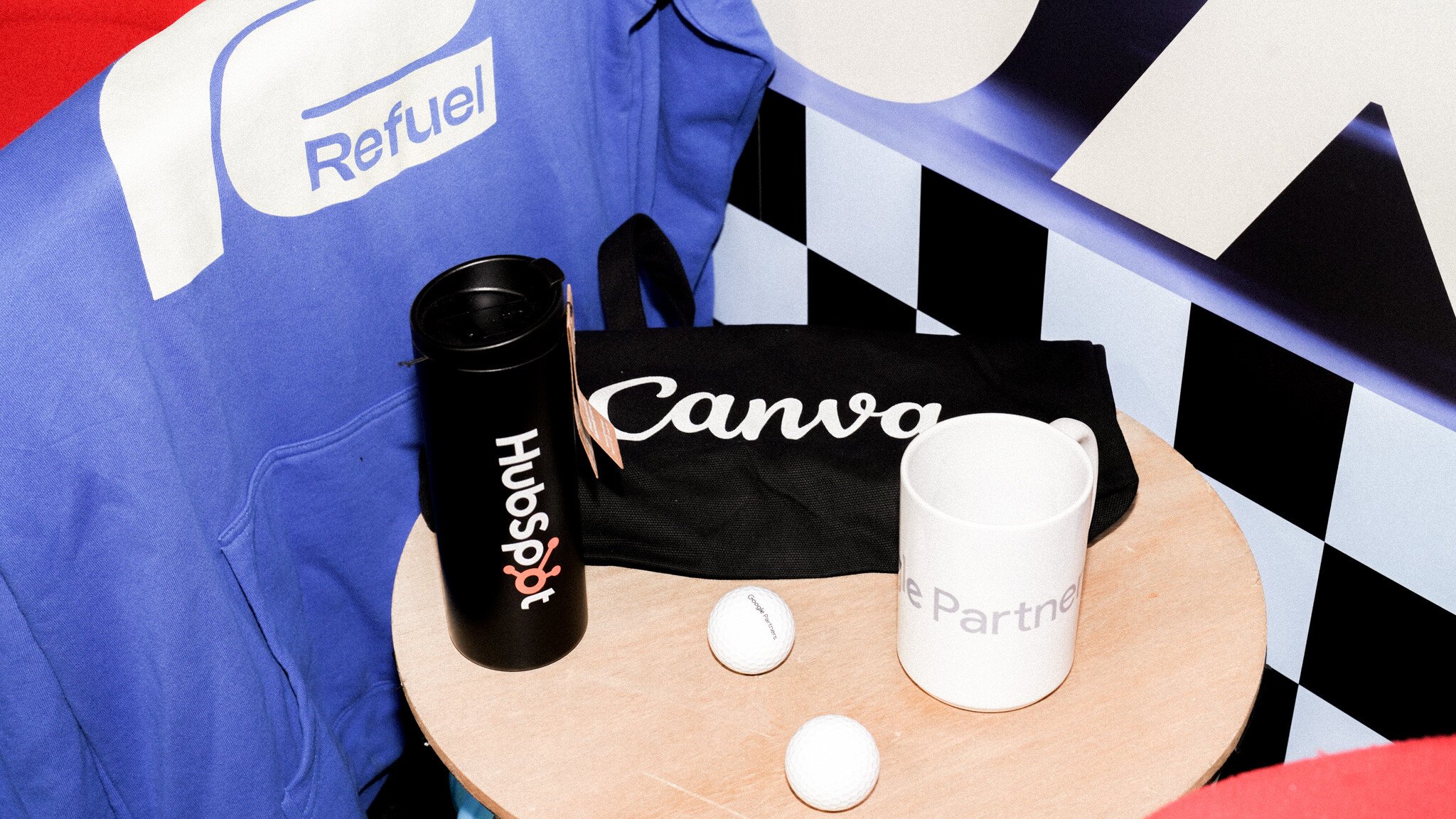 A flat lay photo showing branded merchandise including a black HubSpot water bottle, a black Canva tote bag, and a white 'Partner' mug on a round wooden surface. A blue Refuel banner is visible in the background with a checkered pattern.