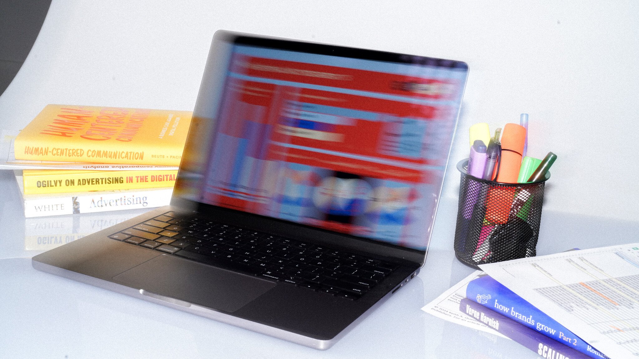 A laptop with motion-blurred screen display, alongside stacked marketing books including 'Ogilvy on Advertising' and 'Human-Centered Communication'. A pen holder with various markers sits to the right.