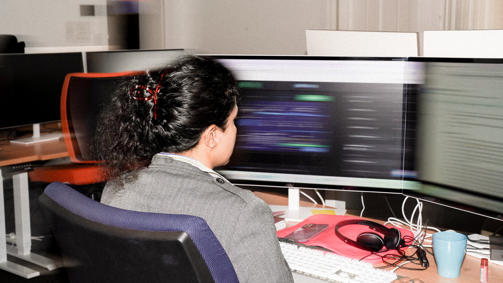 A person viewed from behind working at a dual-monitor setup, with analytics visible on screen, showing SEO (Search Engine Optimisation) work. Their desk includes headphones, a keyboard, and a coffee mug, in a typical digital marketing workspace setting.