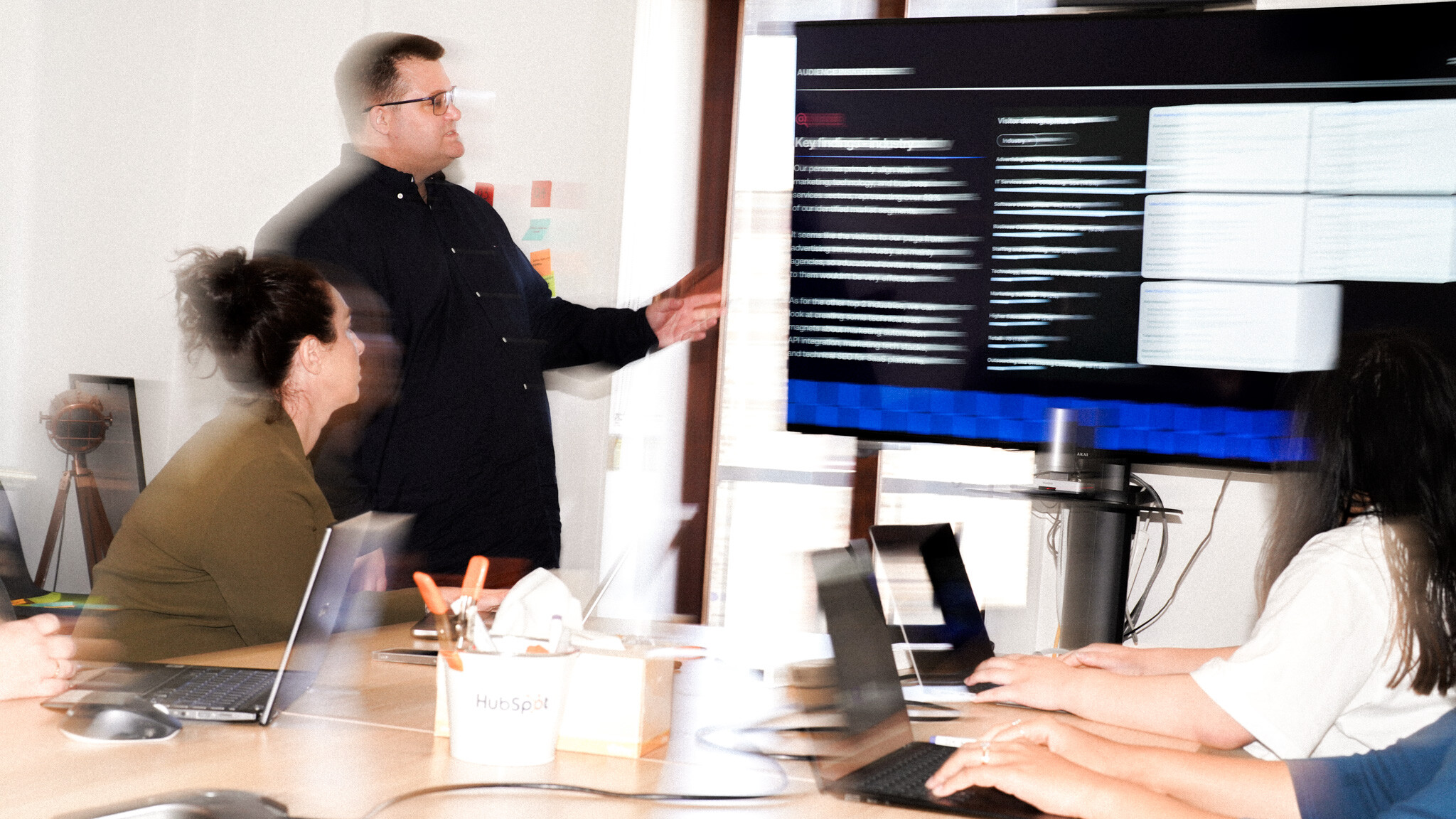 A person in a black shirt standing and gesturing at a wall-mounted screen during what appears to be a business presentation, while others sit at a table with laptops.