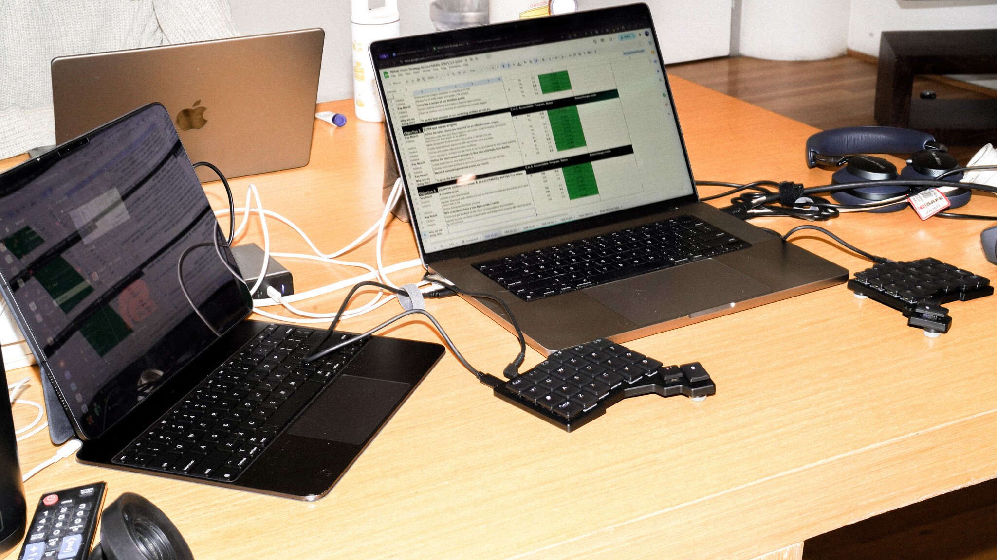 A workspace with two laptops, including a MacBook, displaying spreadsheets. Two split ergonomic keyboards are connected to the laptops. Various cables and peripherals are visible on the wooden desk surface.