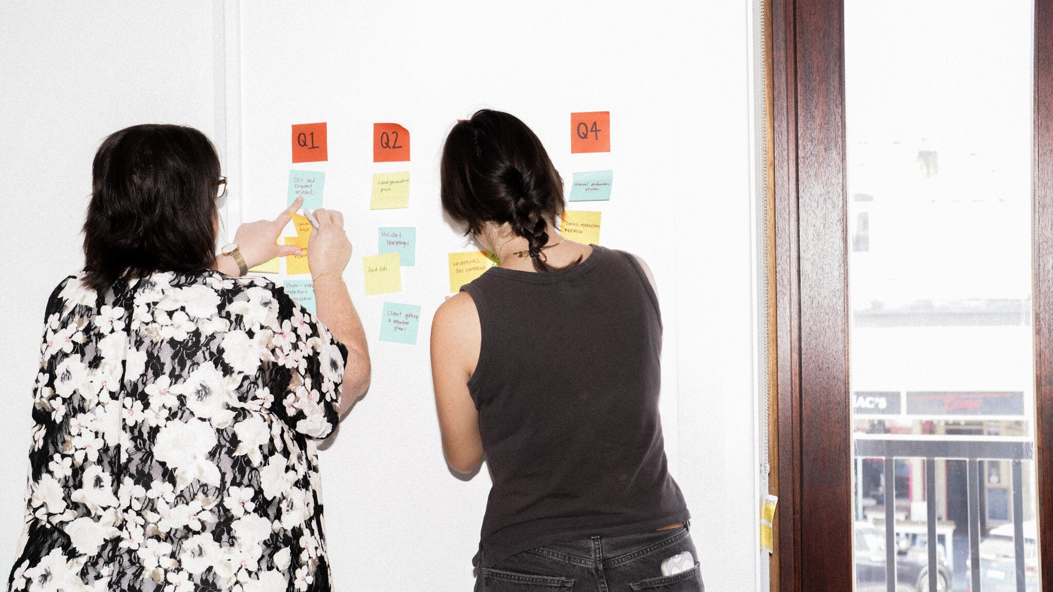 Two people arranging sticky notes on a white wall, organised by quarters (Q1, Q2, Q4). One person wears a black and white floral print top, while the other wears a black sleeveless top.