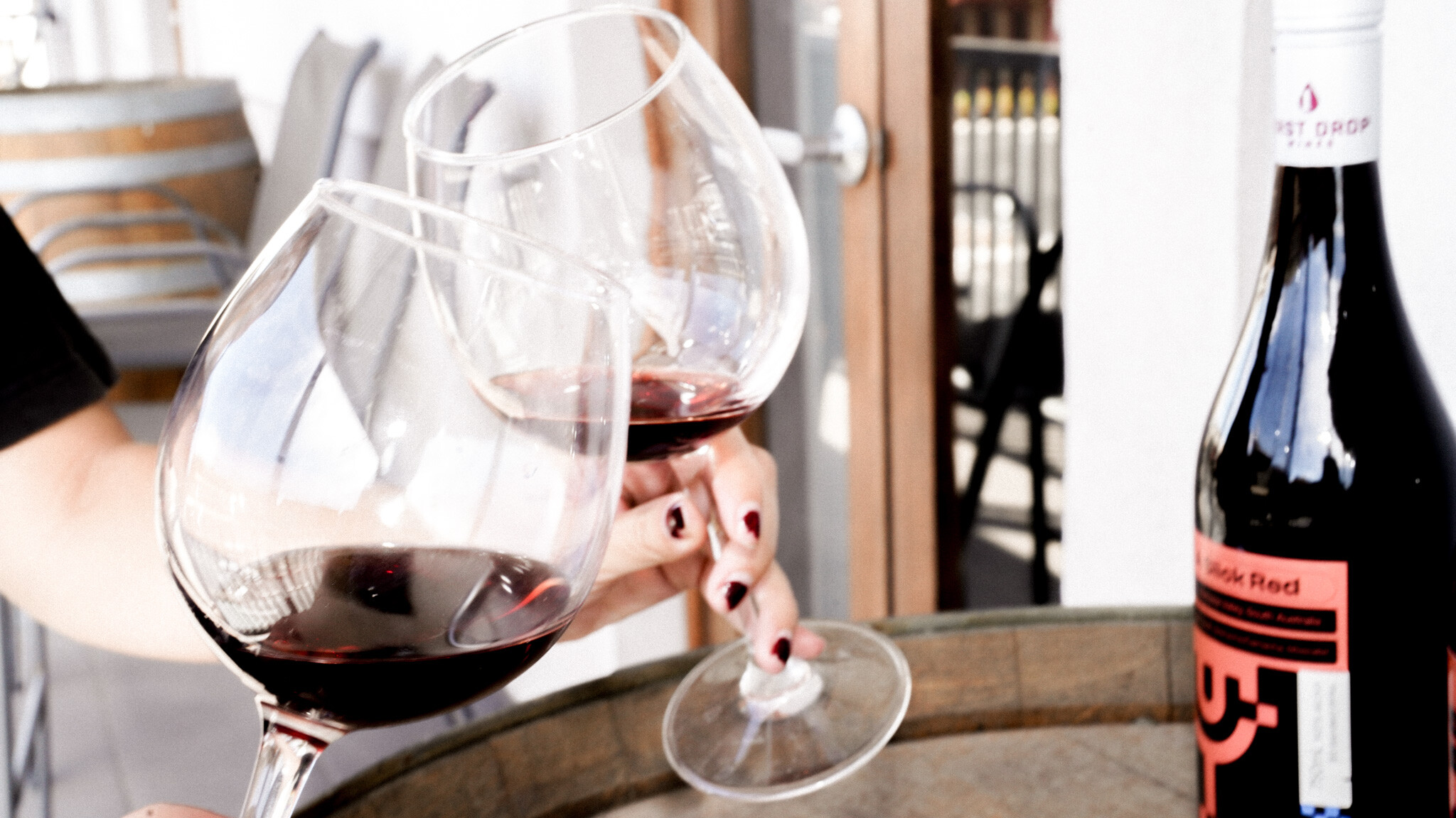 Close-up of two wine glasses clinking together filled with red wine, with a bottle of 'Slick Red' Refuel Creative branded wine visible in the background.