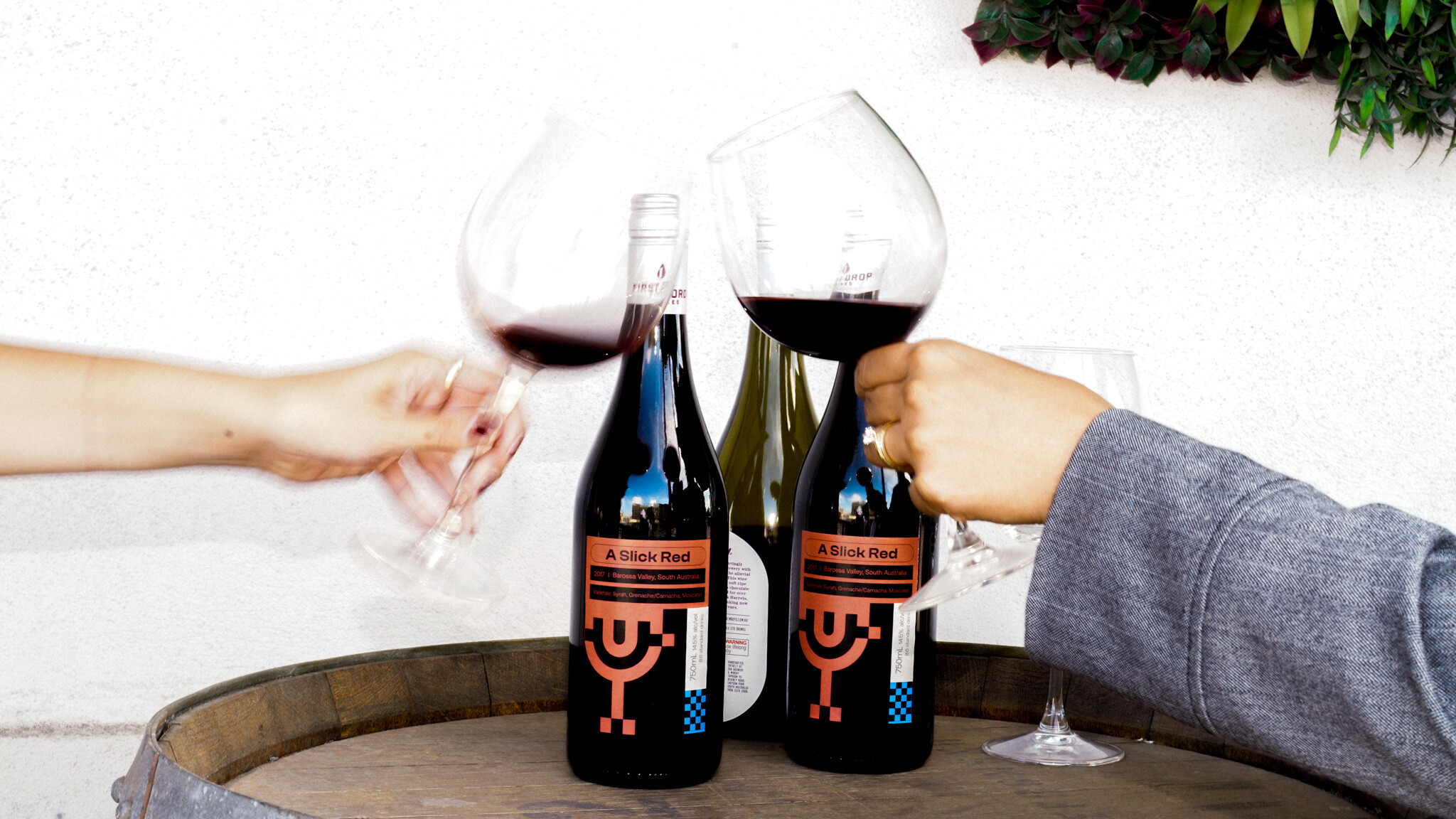 Two hands toasting with wine glasses filled with red wine. Two bottles of 'Slick Red' wine with Refuel-branded labels featuring a minimalist geometric design sit on a wooden barrel.