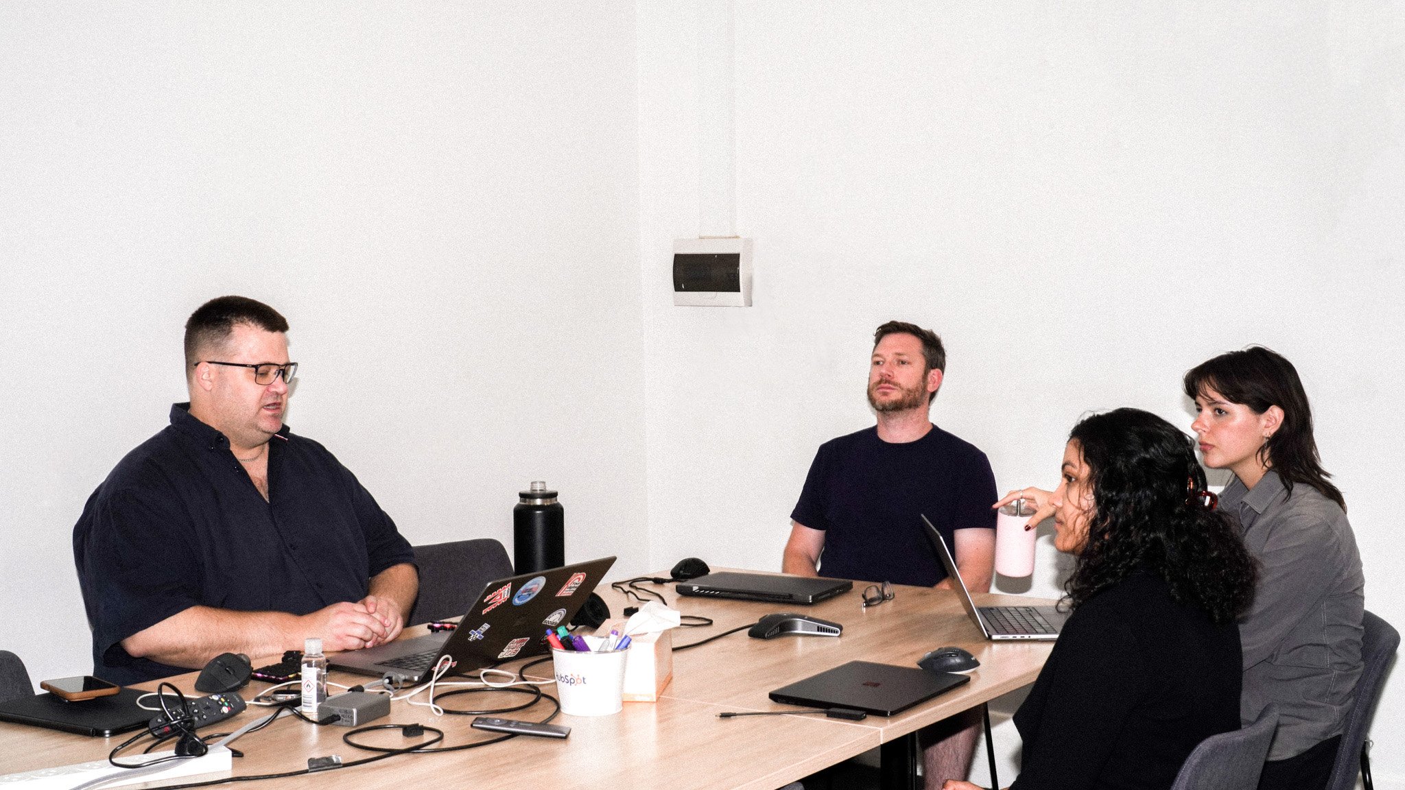 Four people sitting around a wooden conference table having a meeting, with laptops and office supplies scattered on the table.