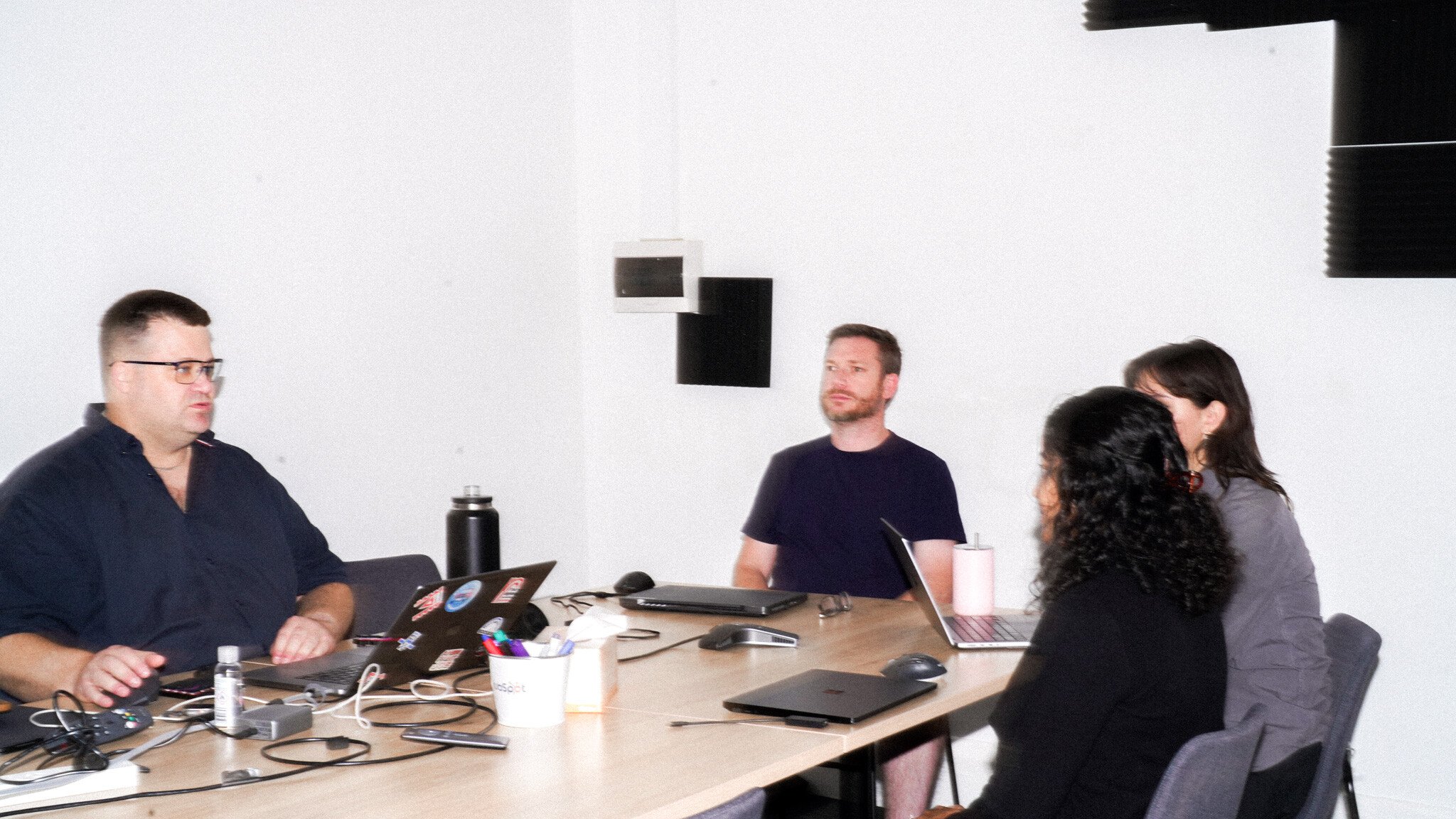 People seated around a wooden conference table having a meeting, with laptops and office supplies scattered on the table. Black geometric shapes are visible on the white wall behind them.