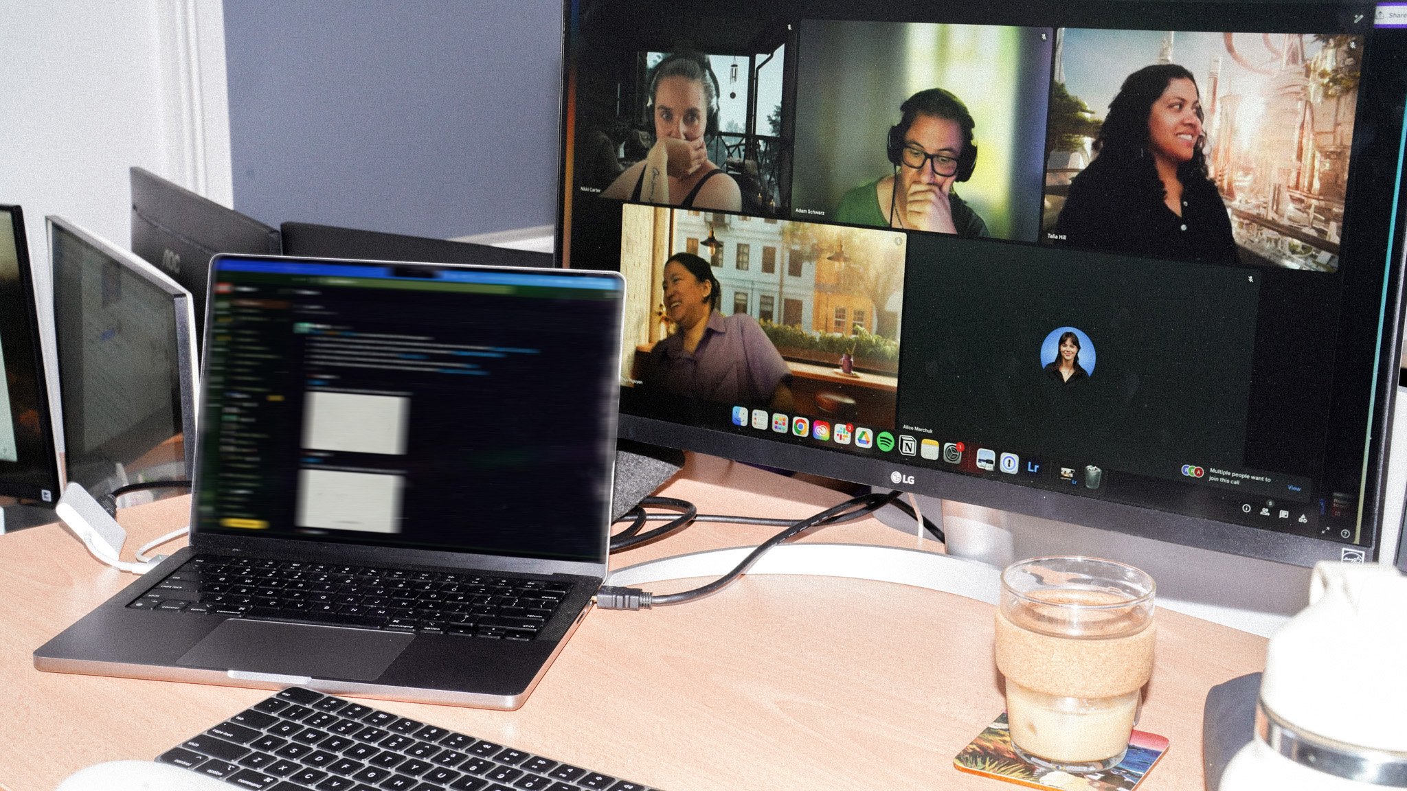 A desk setup showing a laptop and LG monitor displaying a video call with multiple participants. The monitor shows four people in a grid layout against various backgrounds. A glass of coffee sits on the desk next to the devices.