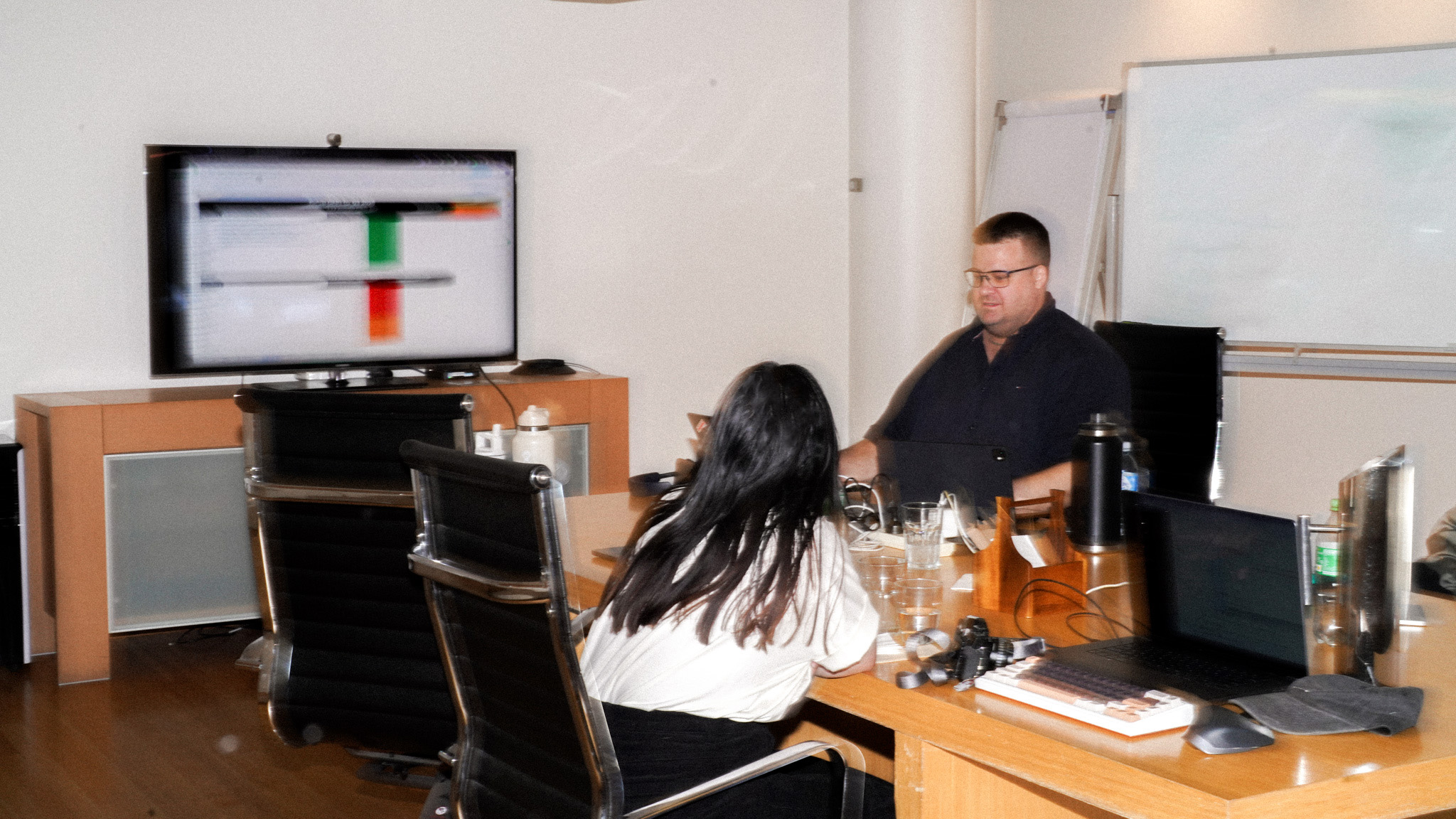 An office setting with two people looking at a screen displaying data visualisations. A TV mounted on the wall shows coloured bars or charts, while laptops and office supplies are scattered on wooden desks.