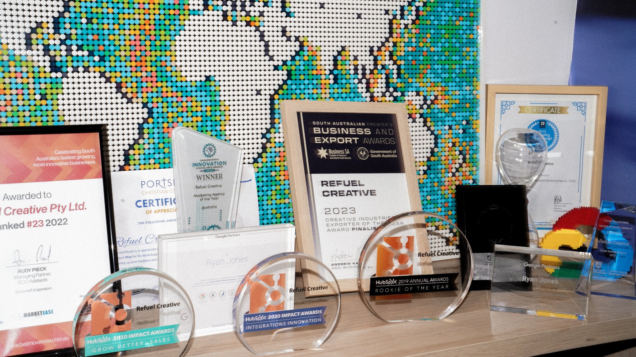 Various business awards and certificates displayed on a shelf, including Refuel Creative awards and trophies, with a colourful dotted world map pattern visible in the background.