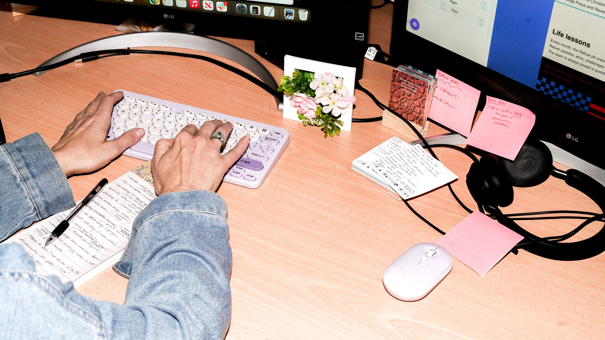 Hands typing on a white keyboard, wearing denim sleeves. The wooden desk has handwritten notes, pink sticky notes, a small floral photo display, headphones, and a white mouse scattered across it.