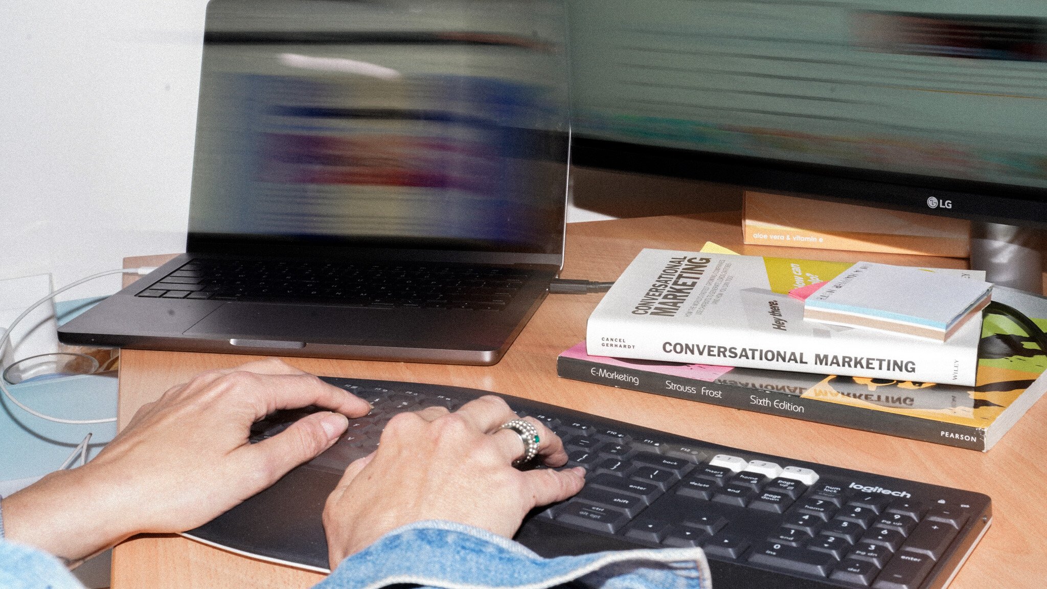 Hands typing on a Logitech keyboard next to a laptop, with marketing textbooks including 'Conversational Marketing' stacked on the wooden desk.