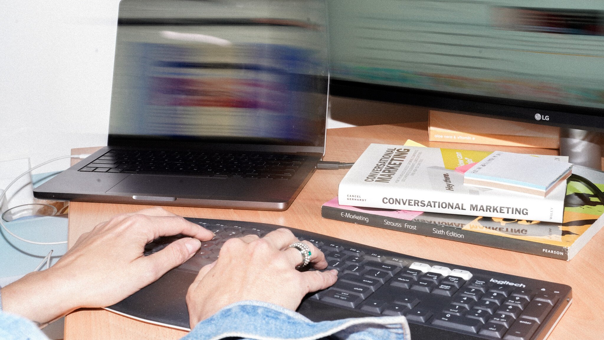 Hands typing on a black keyboard with a laptop and marketing books including 'Conversational Marketing' stacked on a wooden desk. An LG monitor is partially visible in the background.