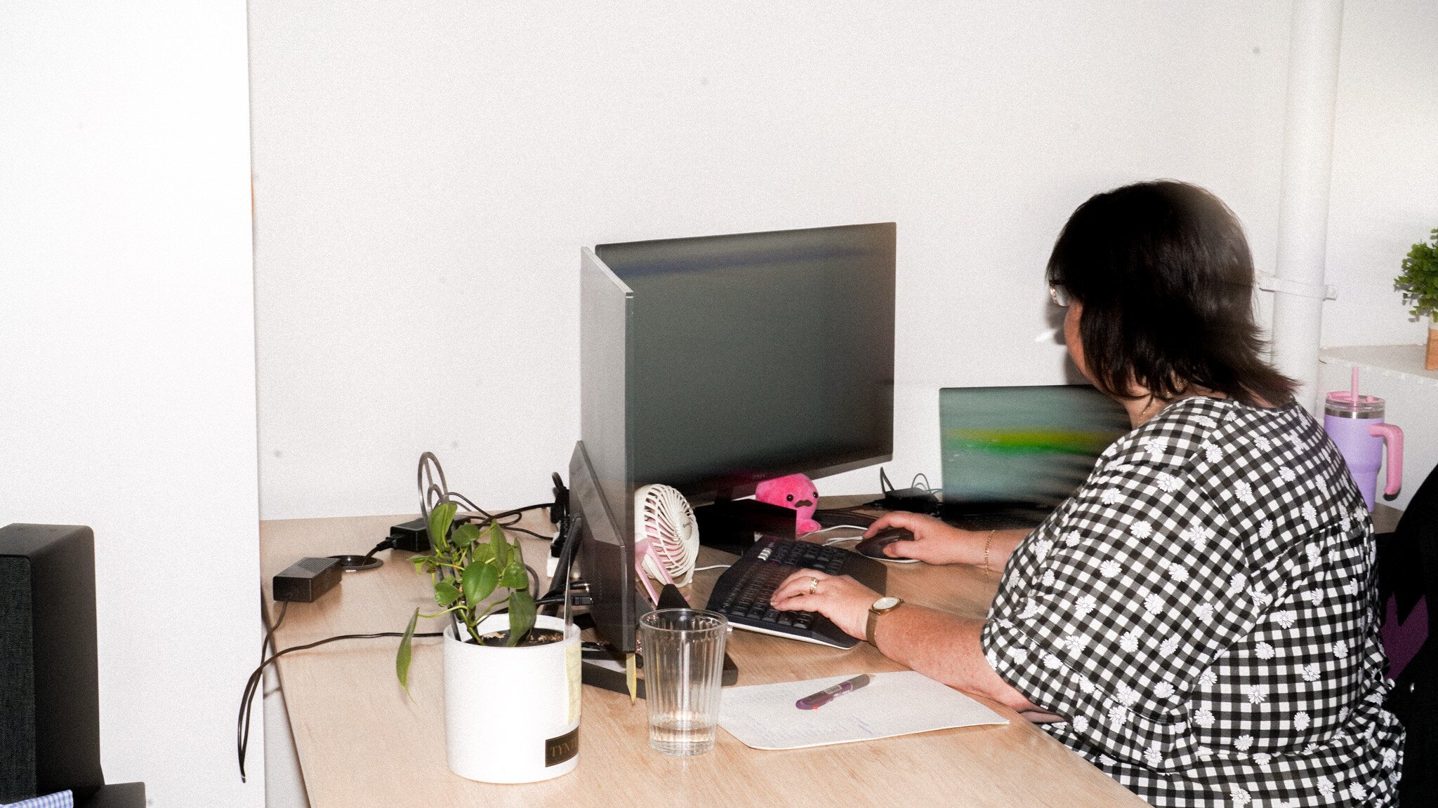 Someone in a checkered shirt working at a desk with dual monitors, a small potted plant, and a mini fan visible. Various cables and office supplies are scattered on the desk surface.