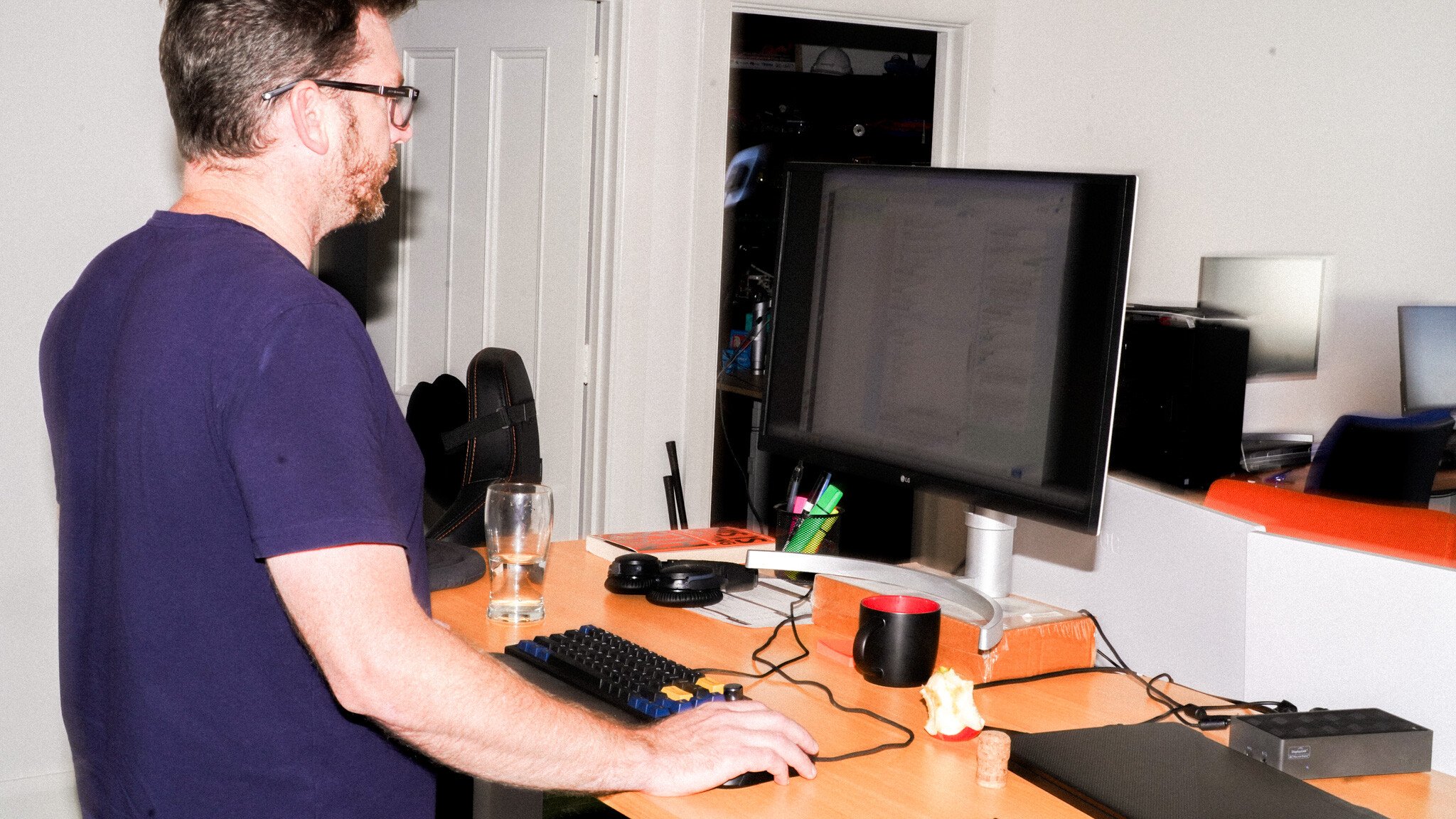 Side view of a person in a purple t-shirt working at a standing desk with a monitor, keyboard, and various office accessories on the wooden surface.