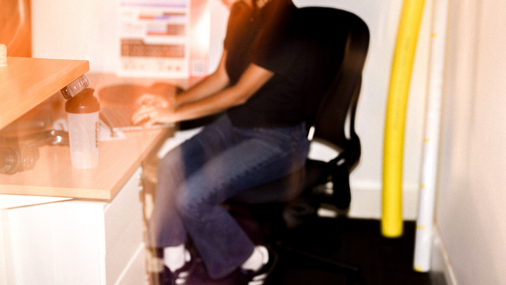 Blurred motion image of someone sitting in an office chair at a desk, with parts of the workspace visible but out of focus.