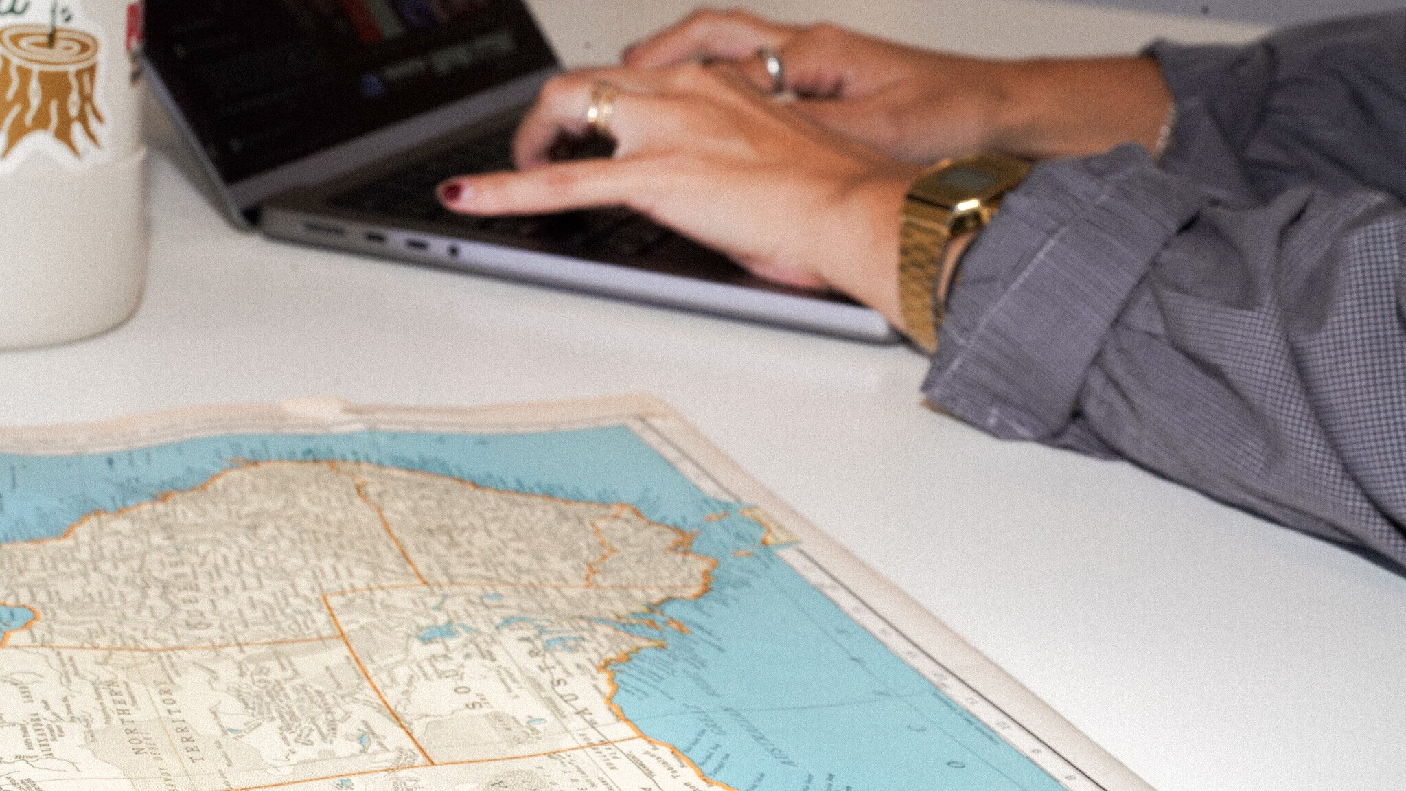 Hands typing on a laptop keyboard, wearing a gold watch, with part of a vintage map of Australia visible on the white desk surface.