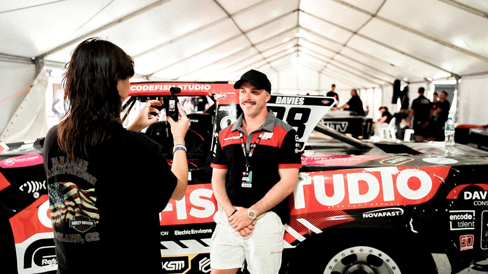 Someone in a black t-shirt filming an interview with a race car driver standing next to a red and black Codefish Studio super truck in a white tent paddock area.