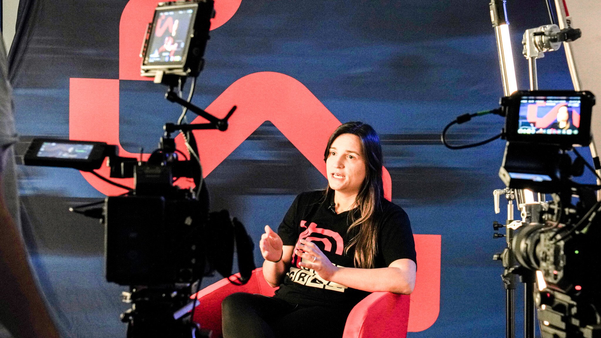 A person in a black t-shirt sitting in a red chair being filmed by multiple video cameras in a studio setting. A red geometric pattern is visible on the blue backdrop wall.