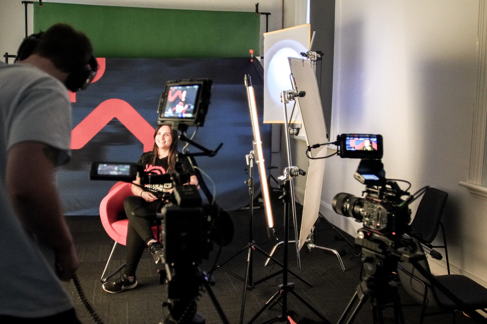 Behind-the-scenes view of a video production set, showing a person in black clothing seated in a red chair being filmed by multiple cameras. Lighting equipment and a blue backdrop with red geometric shapes are visible.