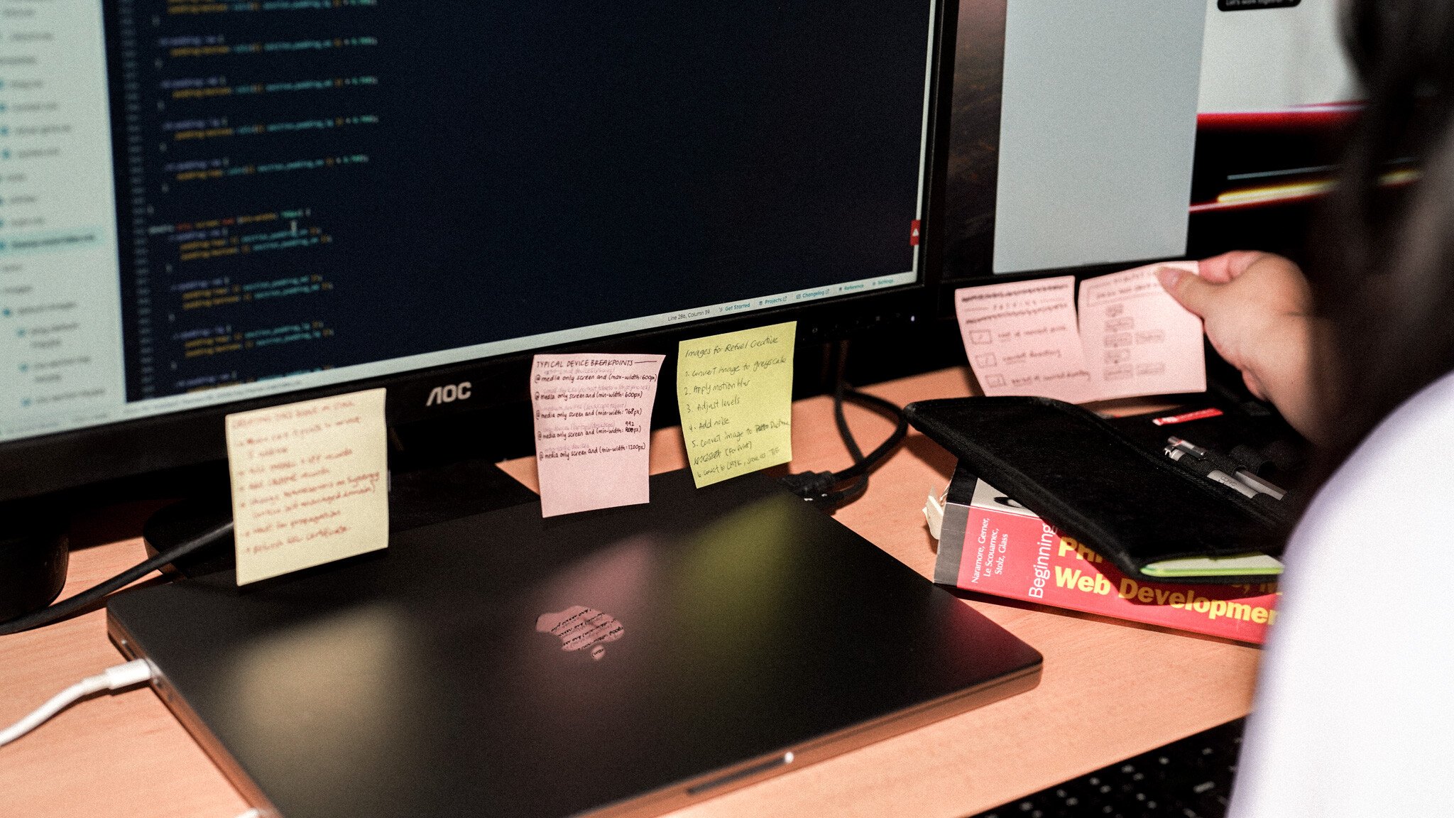 A developer's desk showing an AOC monitor displaying code, with multiple sticky notes attached to its base. A black MacBook and what appears to be a web development book are also visible.