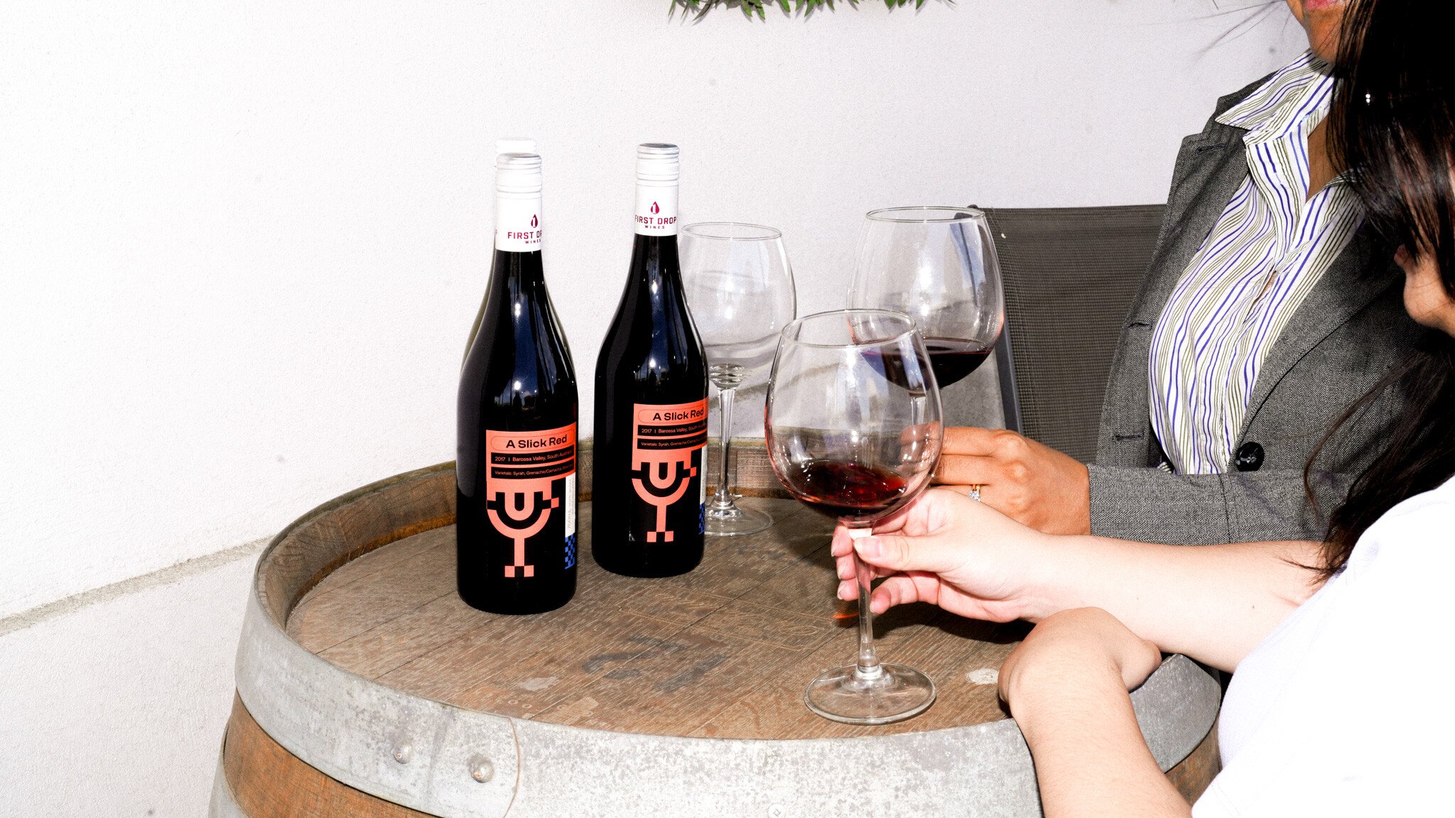 Wine glasses and bottles of 'Slick Red' wine with Refuel branding on a wooden barrel top. A person in a striped shirt and grey jacket is partially visible.