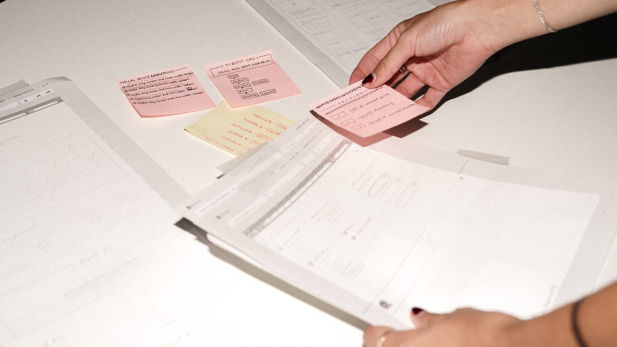 Hands arranging pink and yellow sticky notes alongside paper wireframes, showing a web design planning process with sketched layouts and interface notes.