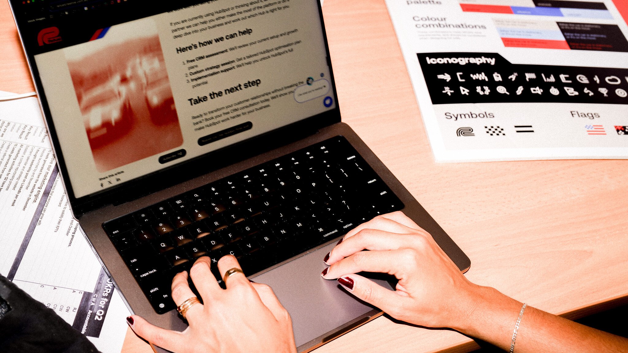 Hands typing on a laptop keyboard, with rings visible on the fingers. A typography and symbols reference guide is visible on the desk alongside other documents.