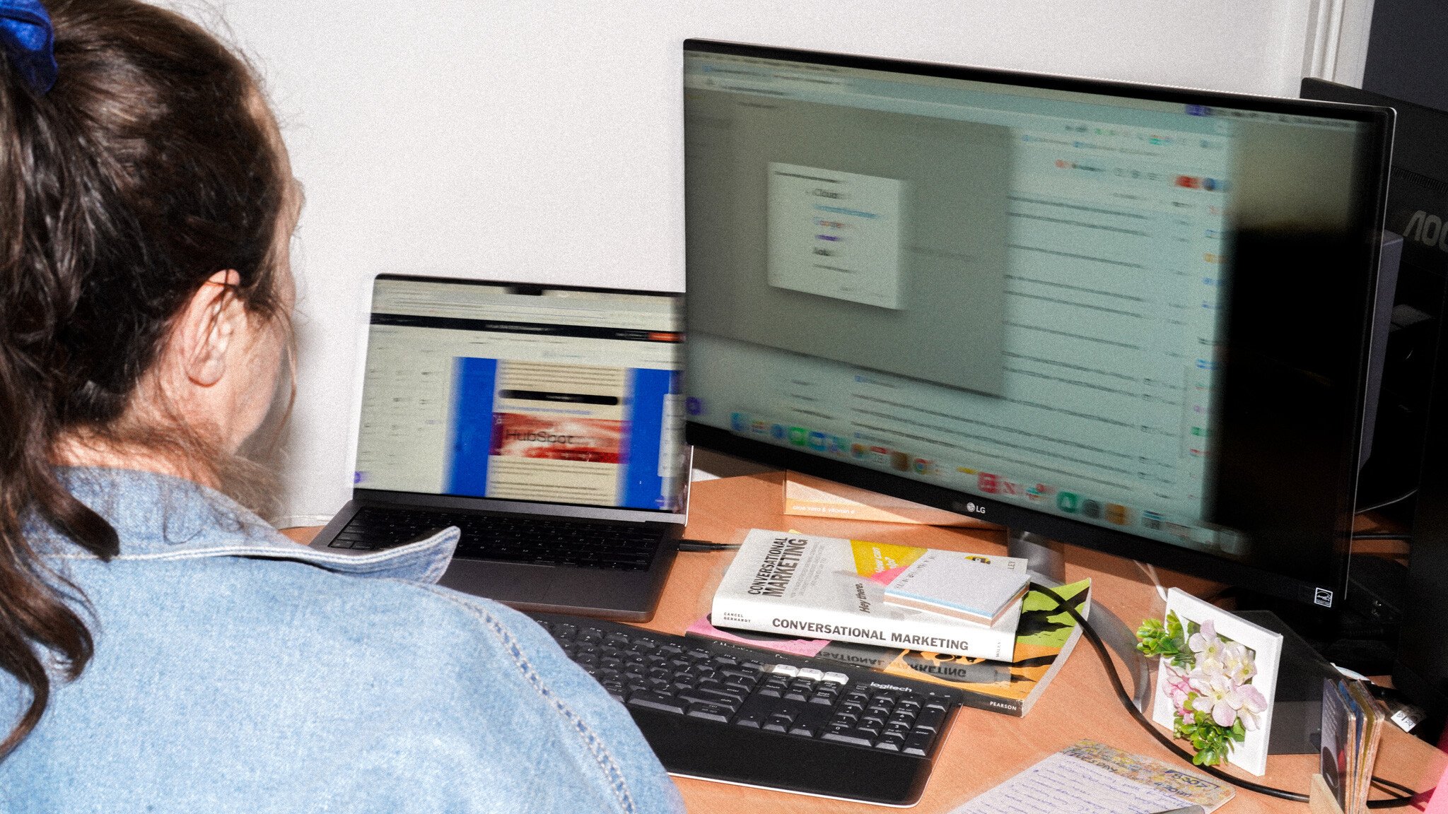 A person in a denim jacket working at a desk with two monitors, displaying email marketing campaign interfaces. A book titled 'Conversational Marketing' and other materials are visible on the desktop.