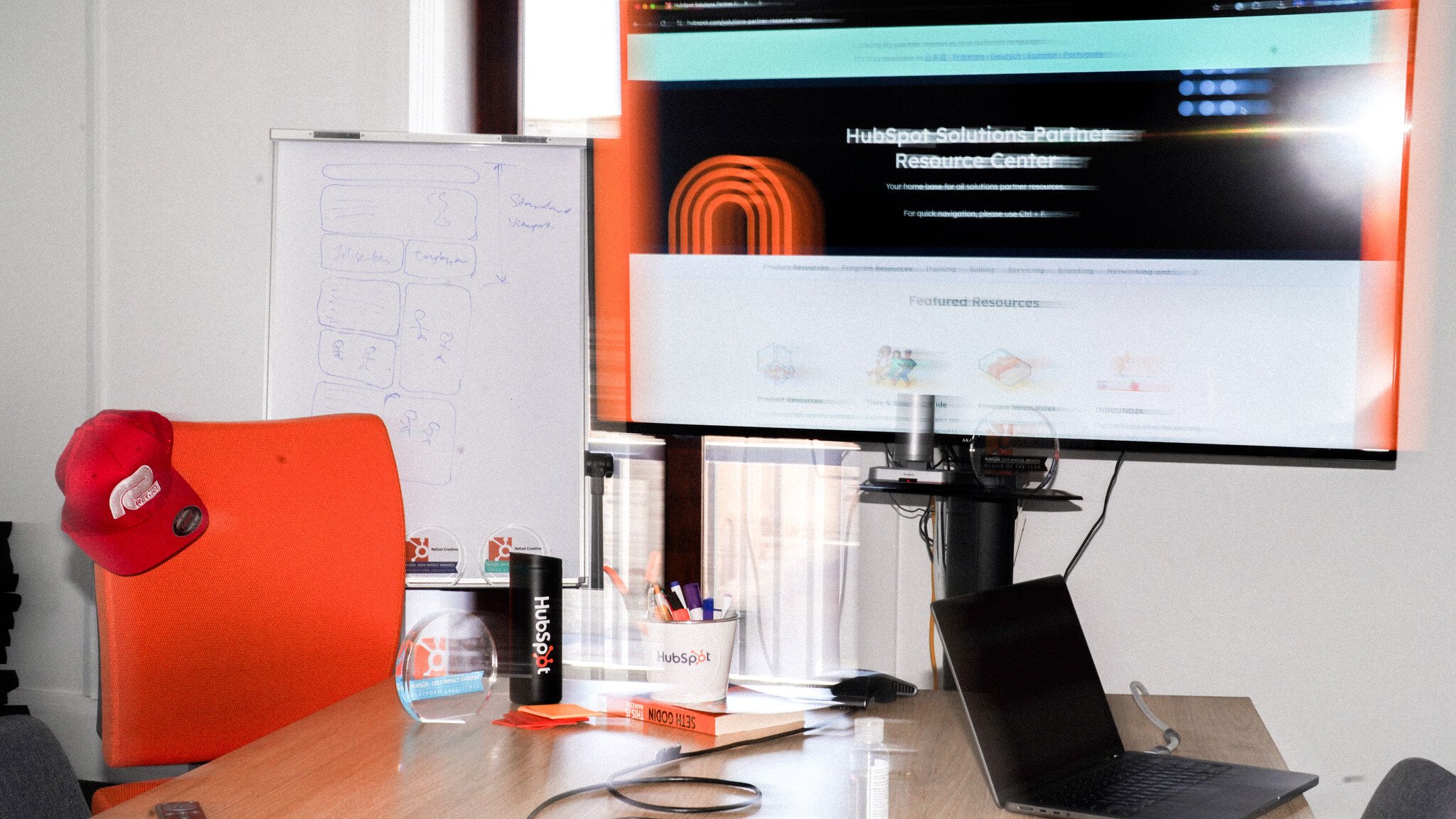 A workspace showing HubSpot's Solutions Partner Resource Center on a monitor, with HubSpot branded merchandise including a water bottle and pen holder. An orange office chair, red Refuel Creative baseball cap, and whiteboard with workflow diagrams are visible in the background.